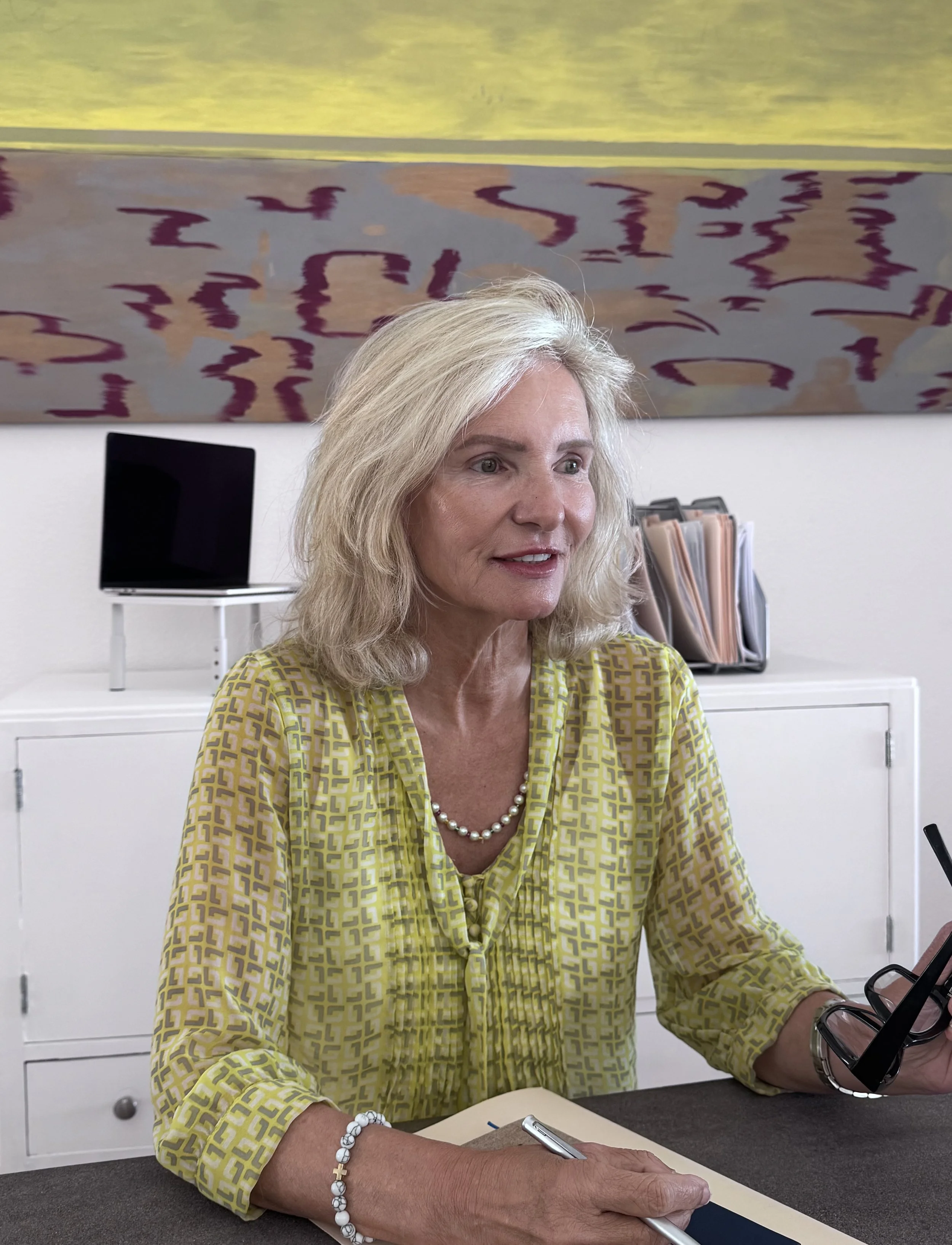 Dr. Valerie Leclercq sitting at her desk in her therapy office in Henderson, Nevada, holding glasses and a pen while speaking with a client.