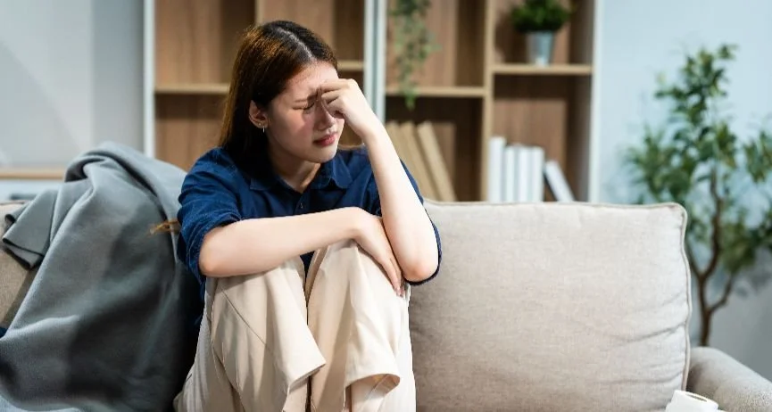 A woman sitting on a couch inside of a room. Her eyes are closed and her hand is touching her face.