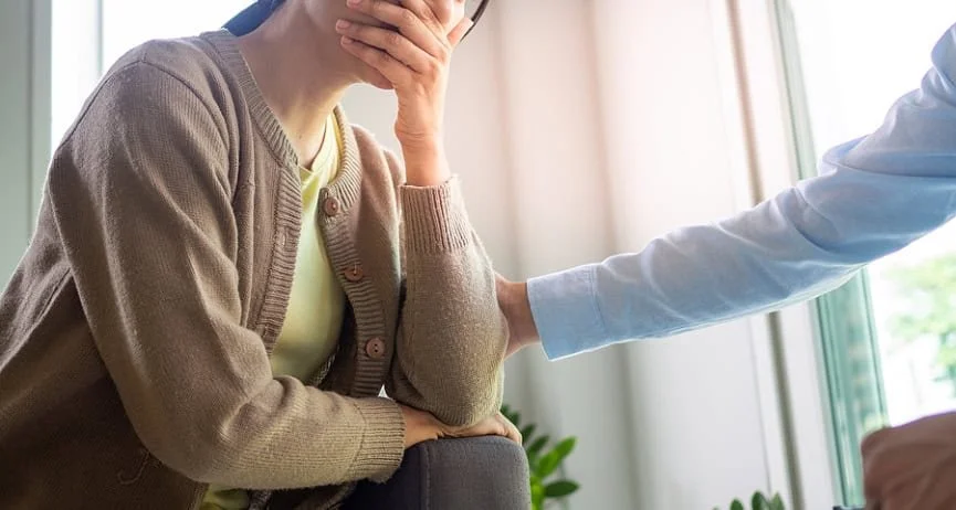 Woman holds hand to her mouth, upset, as someone puts comforting hand on her arm