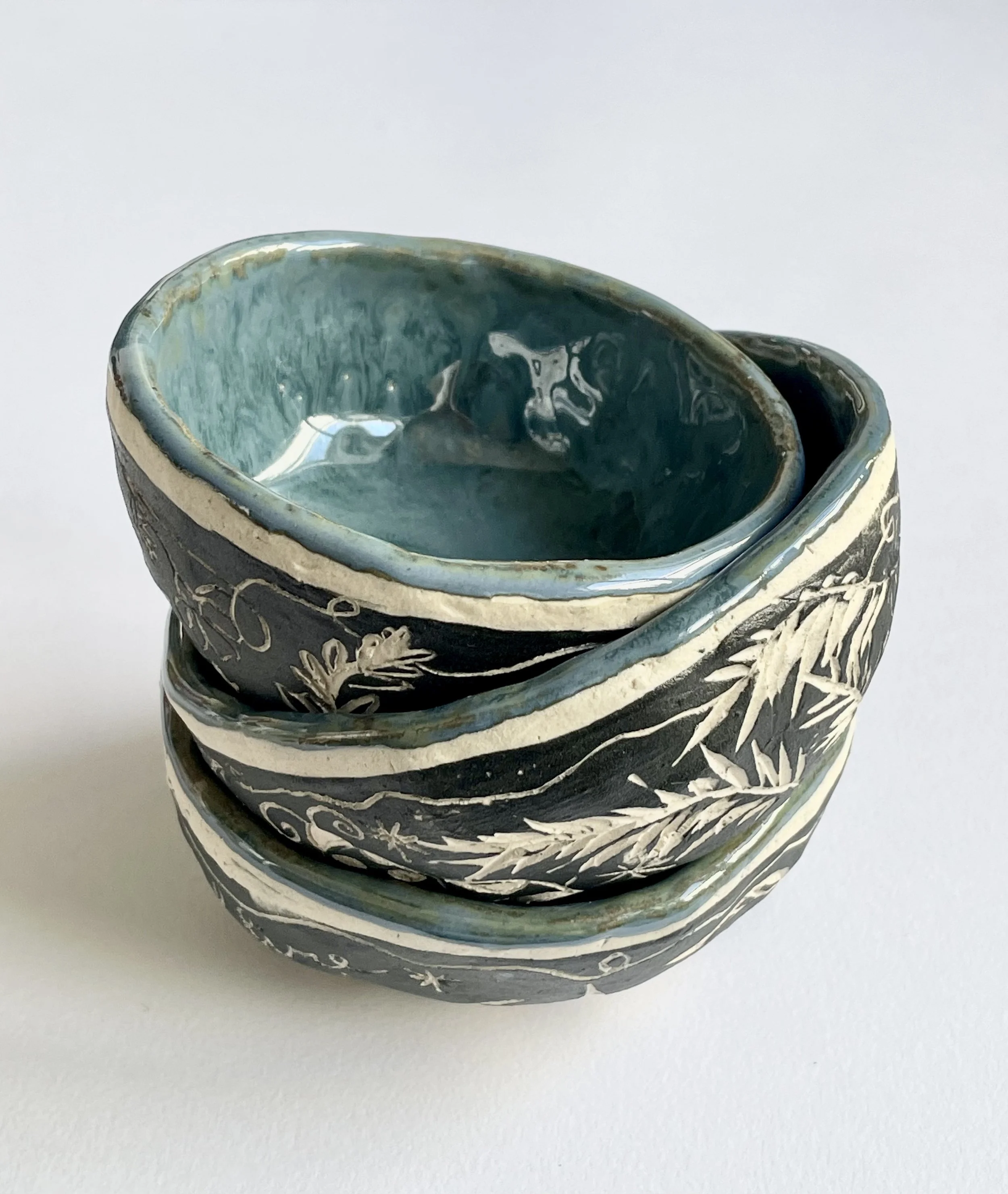 Stacked ceramic bowls with black exterior decorated with white botanical patterns and a teal interior, on a white background.
