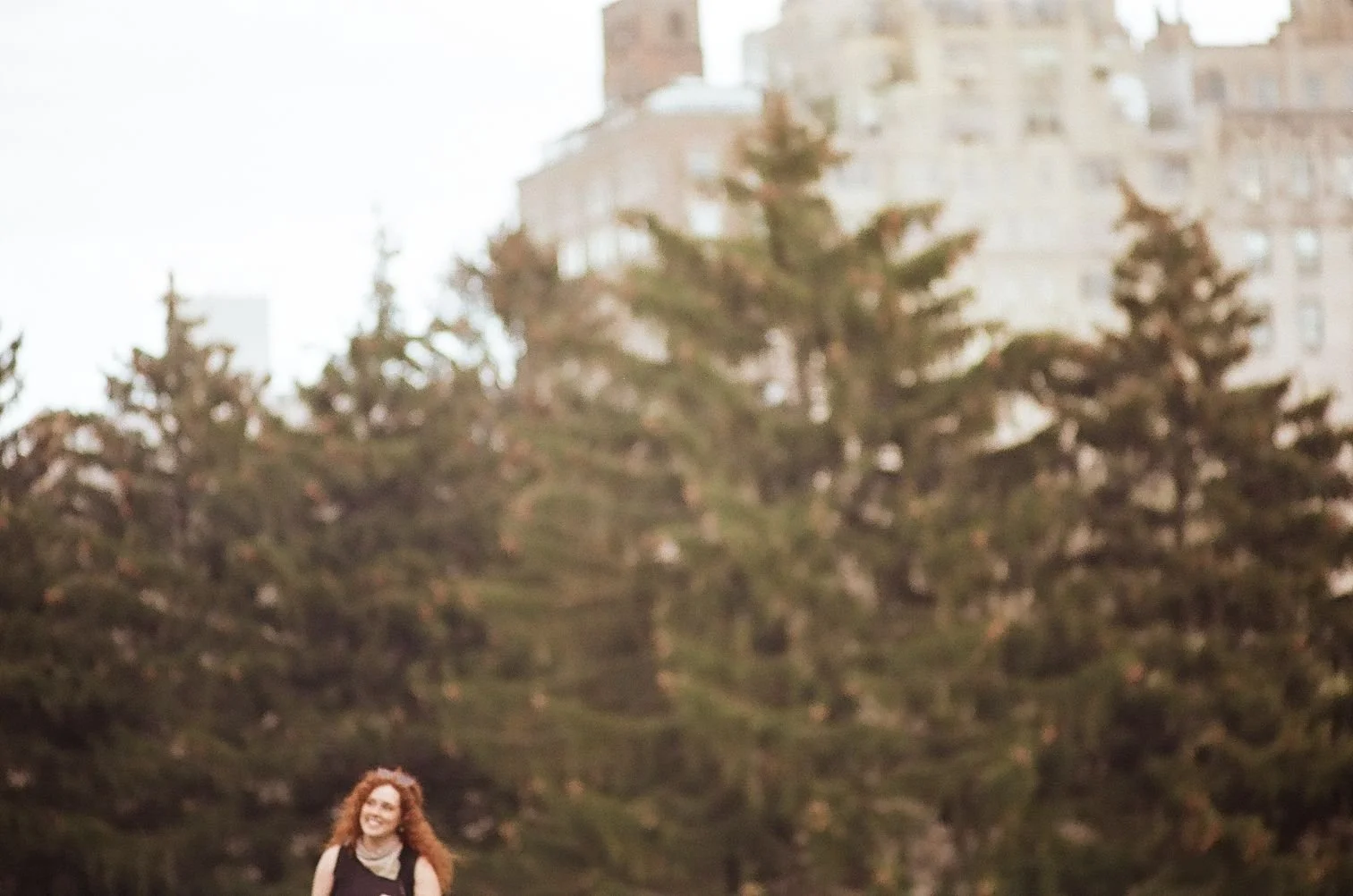 A woman with curly red hair smiling outdoors, with trees and a building in the background.