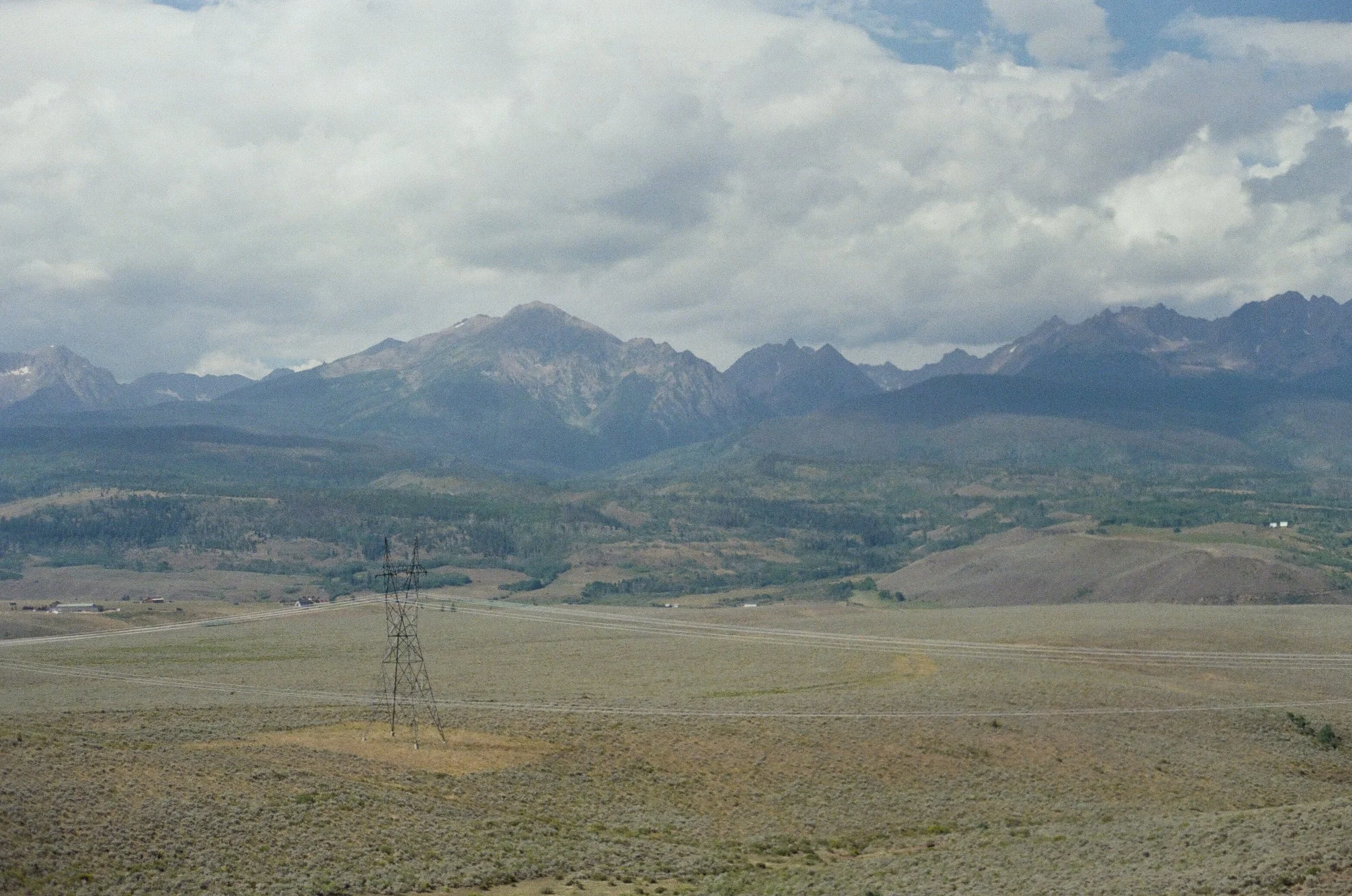 A landscape with rolling hills, power lines, and a mountain range in the distance under a cloudy sky.