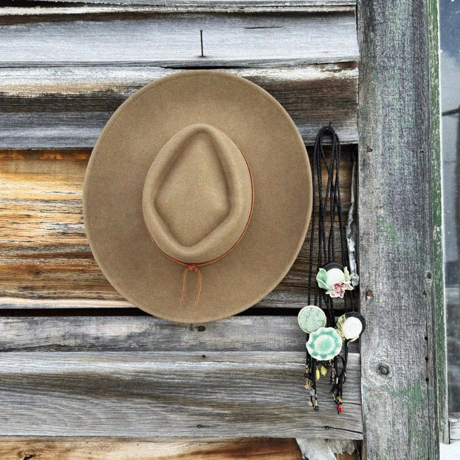 A tan cowboy hat hanging on a weathered wooden wall accompanied by necklaces with floral and natural motifs.