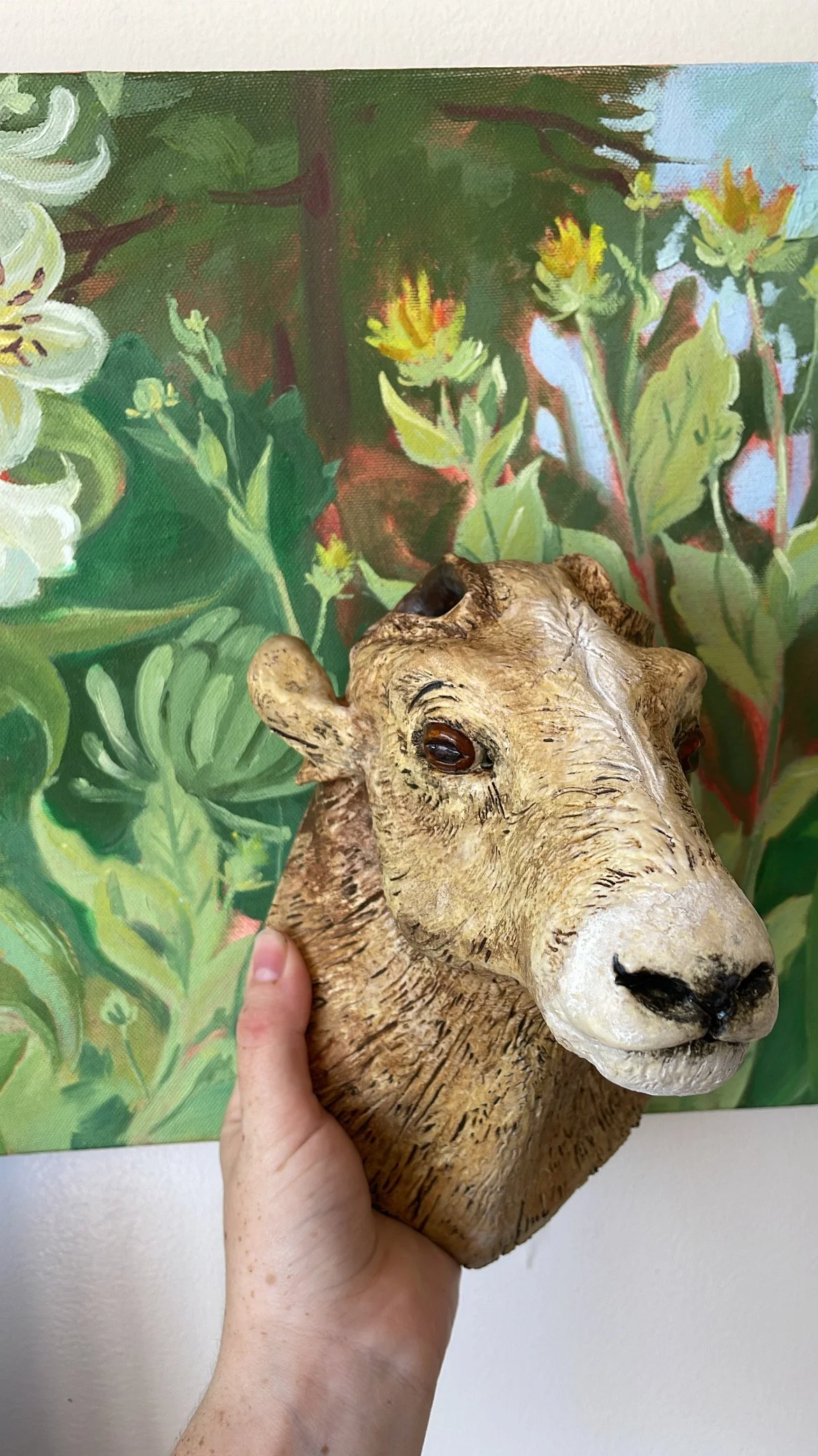 A person holding a realistic-looking animal head sculpture resembling a gnu or wildebeest, with a painted floral background.