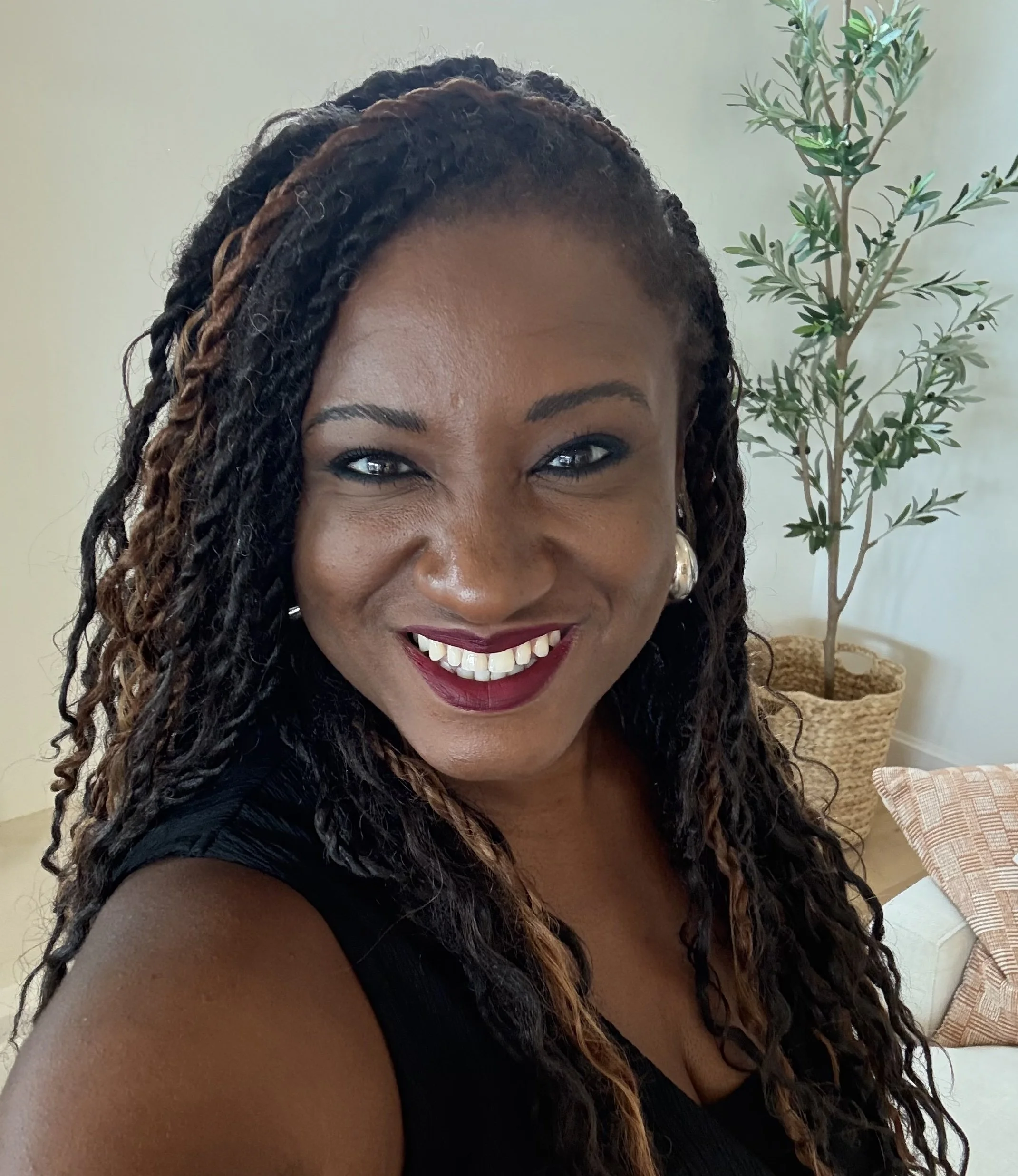 A smiling woman with short curly hair, wearing hoop earrings and a black zip-up top, posing against a light-colored background.