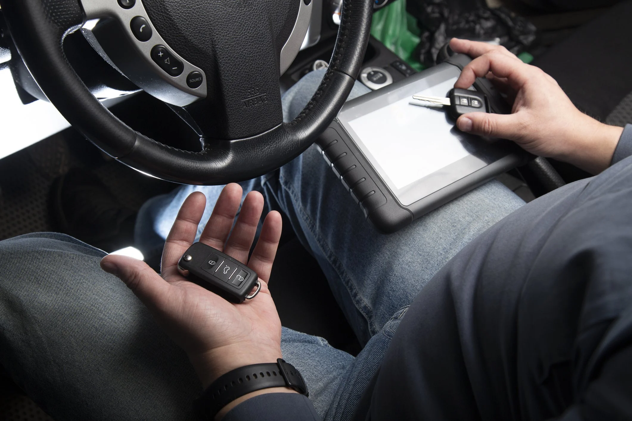 Person sitting in the driver's seat of a vehicle, holding a car key in one hand and a diagnostic scanner with a keyboard and screen in the other. The steering wheel and part of the dashboard are visible.