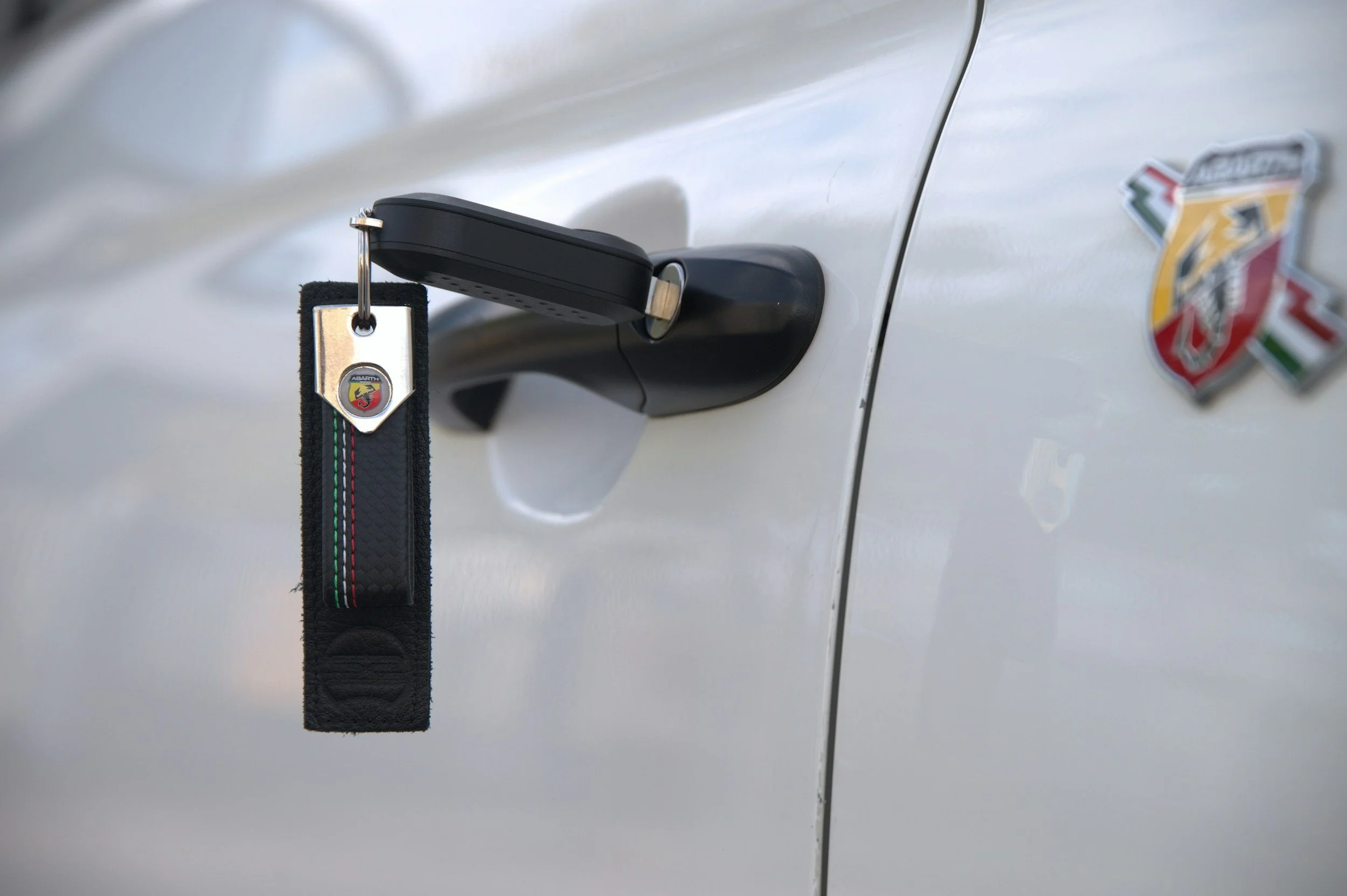 A white car door with a black car key fob hanging from the door handle, attached to a keychain with an Abarth logo, and a badge on the car featuring the Abarth logo with the Italian flag colors.