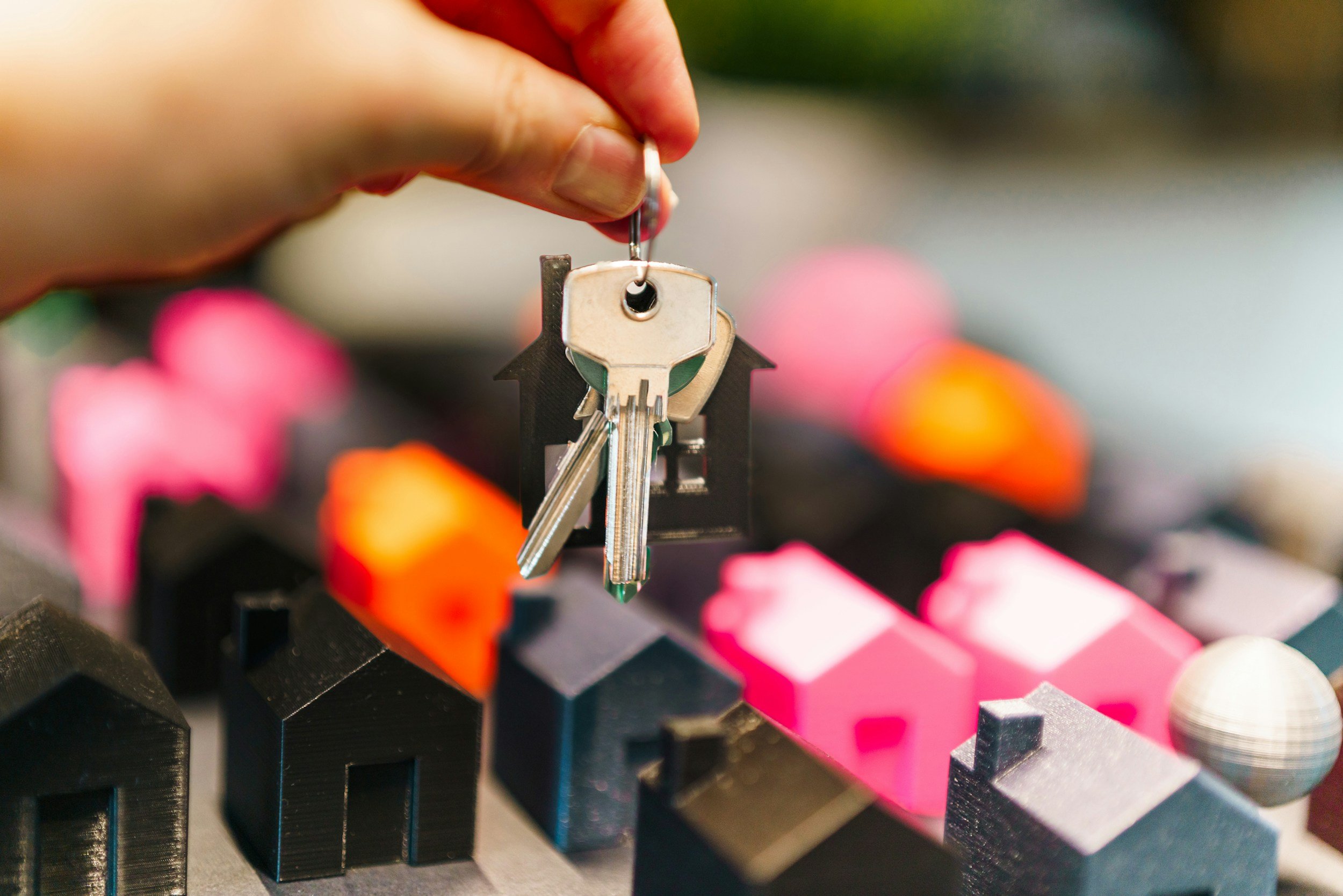 A hand holding a keychain with a house-shaped figure, with colorful miniature houses in the background.