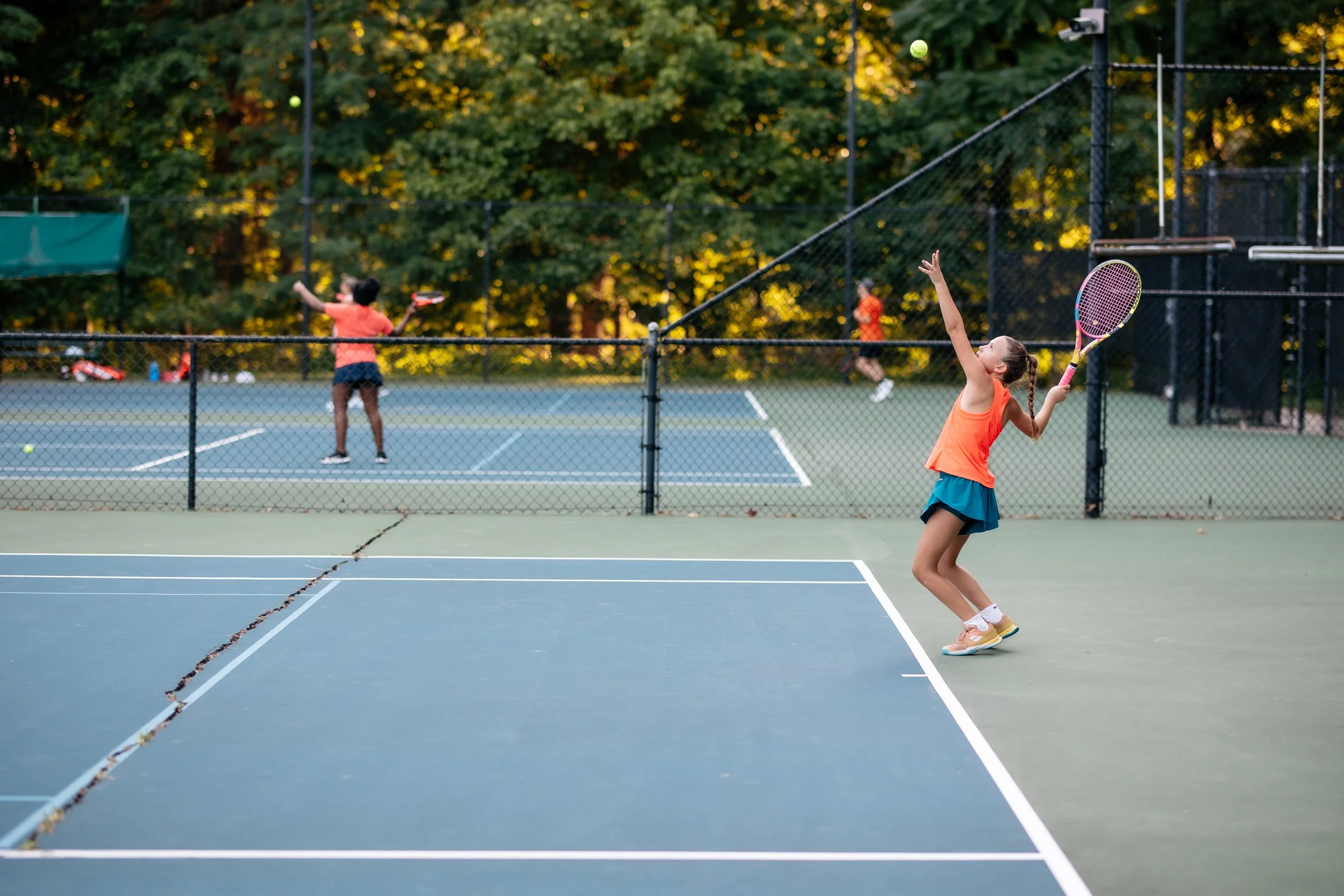 Young girl playing tennis on an outdoor court, preparing to hit a tennis ball with a pink racket. Two other children are playing on a different court in the background, surrounded by greenery.