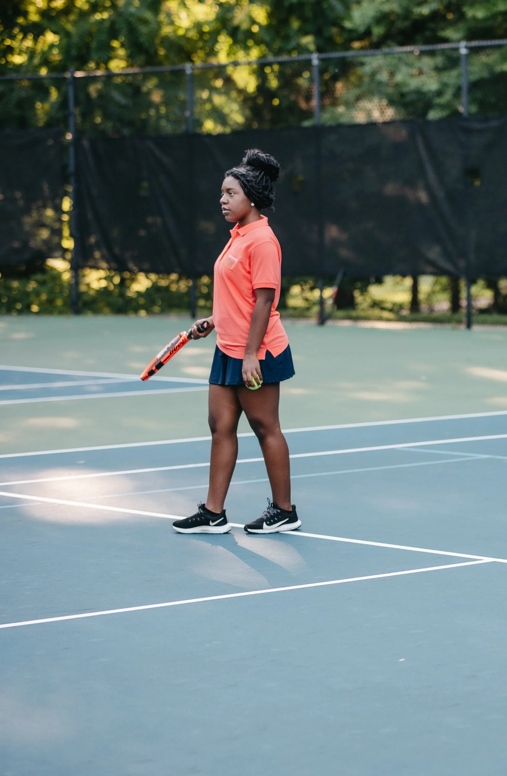 A woman standing on a tennis court holding a tennis racket, wearing an orange polo shirt, navy skirt, and black athletic shoes, with a tennis ball in her hand.