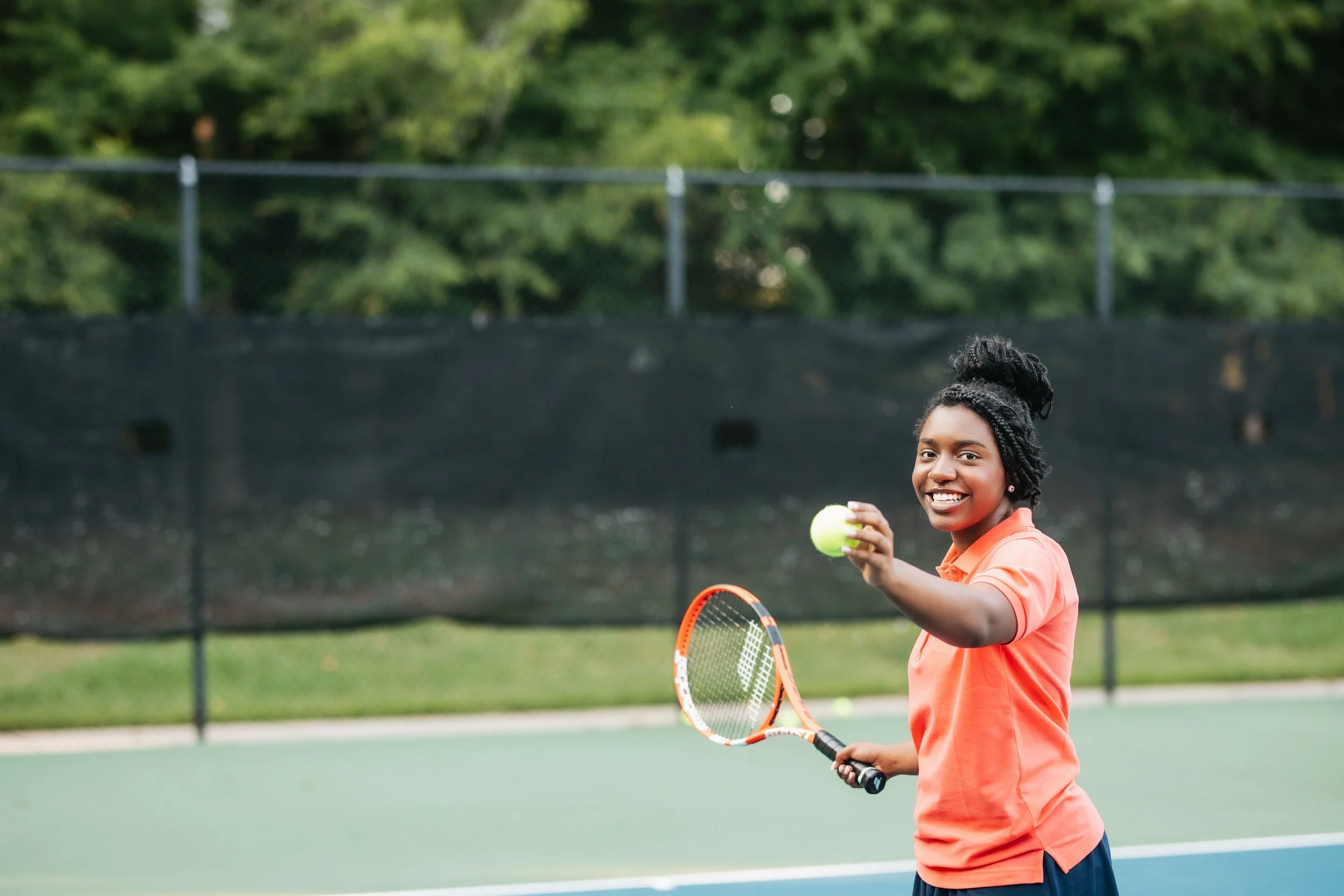 Young woman smiling on tennis court holding tennis ball and racket, dressed in orange polo shirt and navy shorts, with a green tennis court and black windscreen in the background.