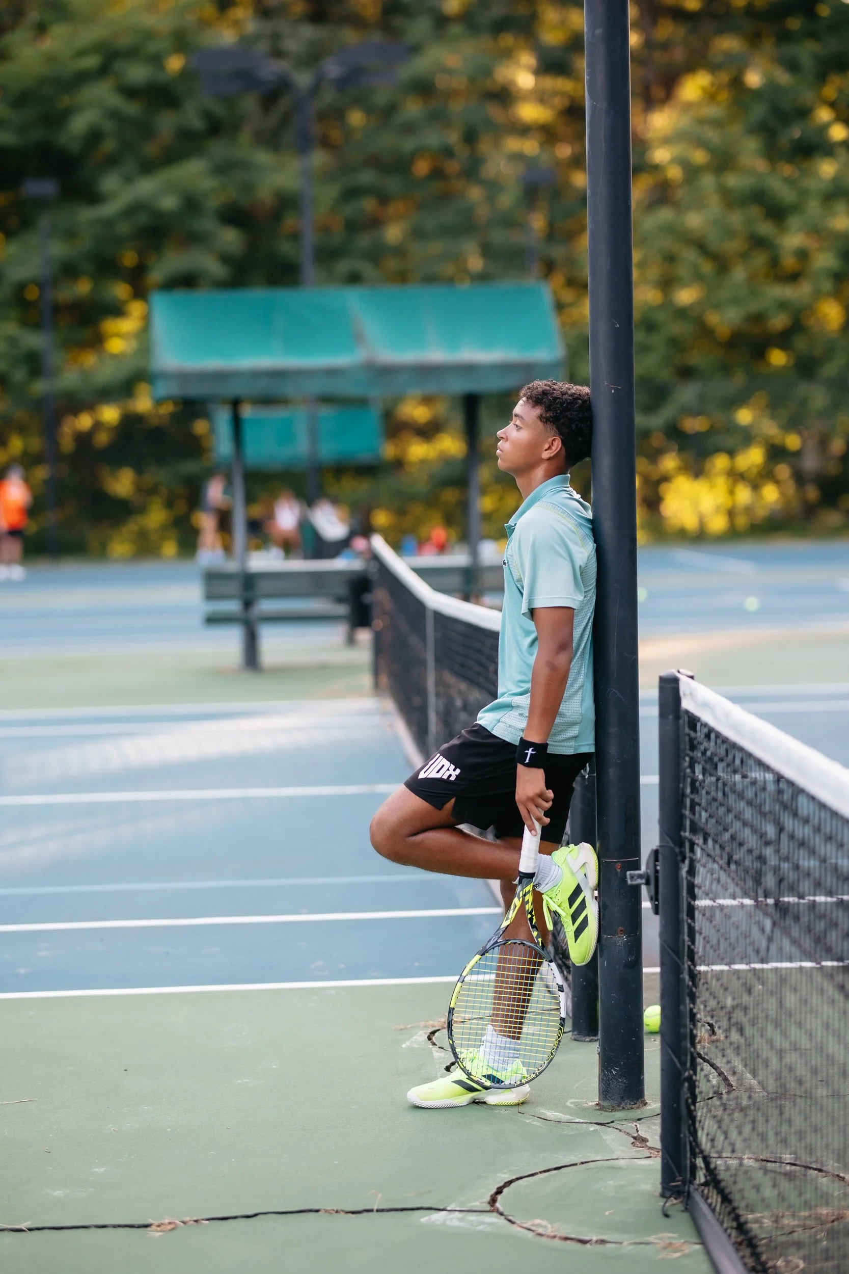 Young male tennis player standing against a tennis net post with his eyes closed, holding a tennis racket, on an outdoor tennis court surrounded by trees.