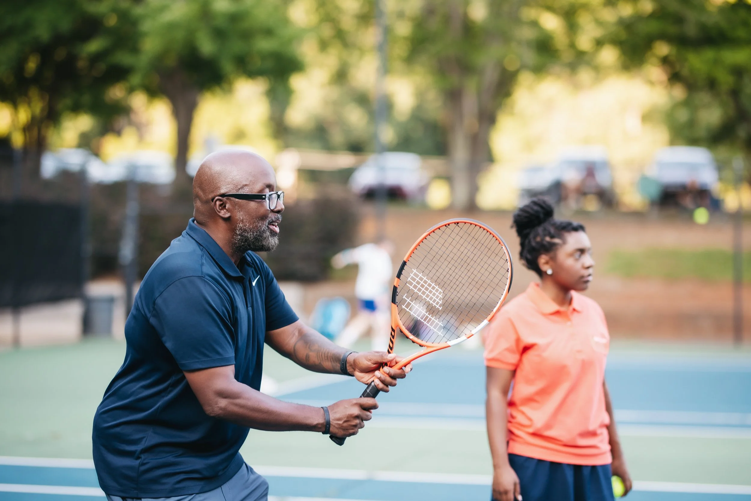 Engaged in coaching tennis, Denard McLendon holds a tennis racket and stands near a woman in orange on a tennis court surrounded by green and the fading light of day.
