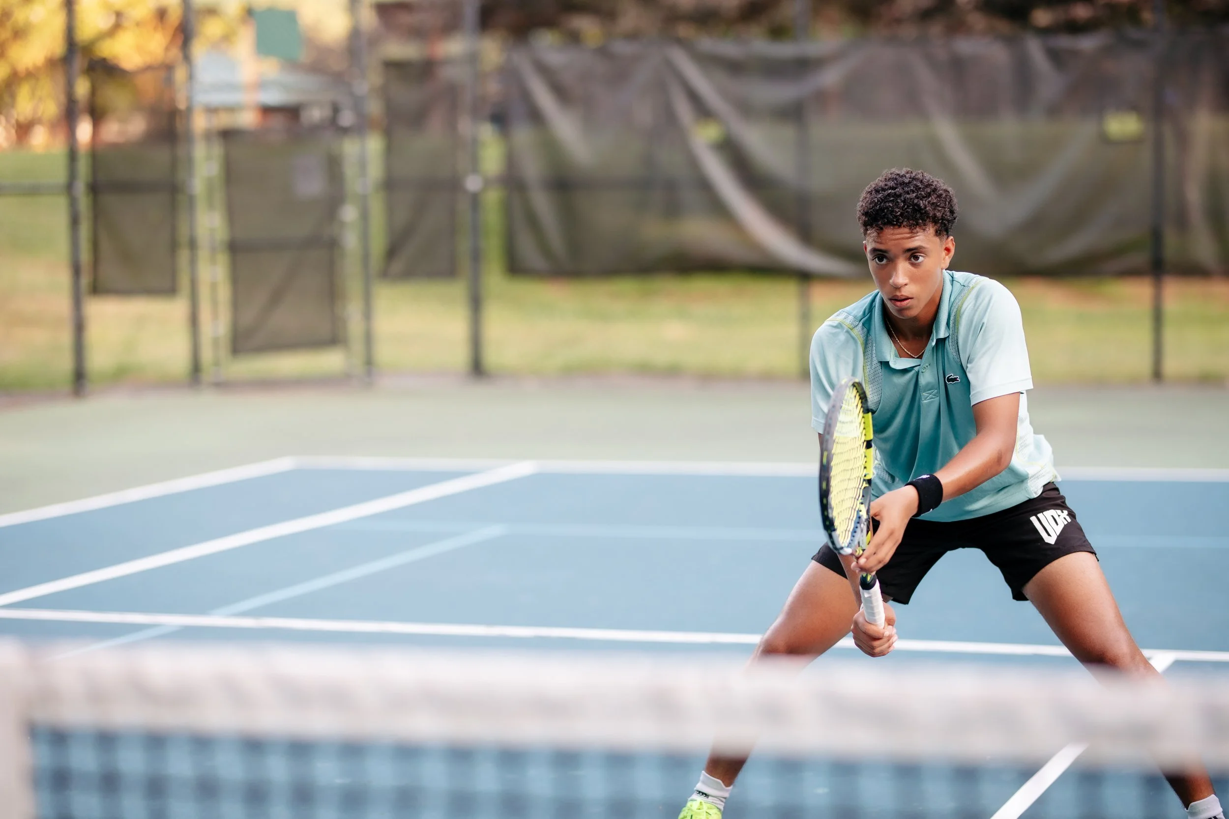 A young male tennis player in a light blue shirt and black shorts is preparing to hit a shot on an outdoor tennis court with trees and a black fence in the background.