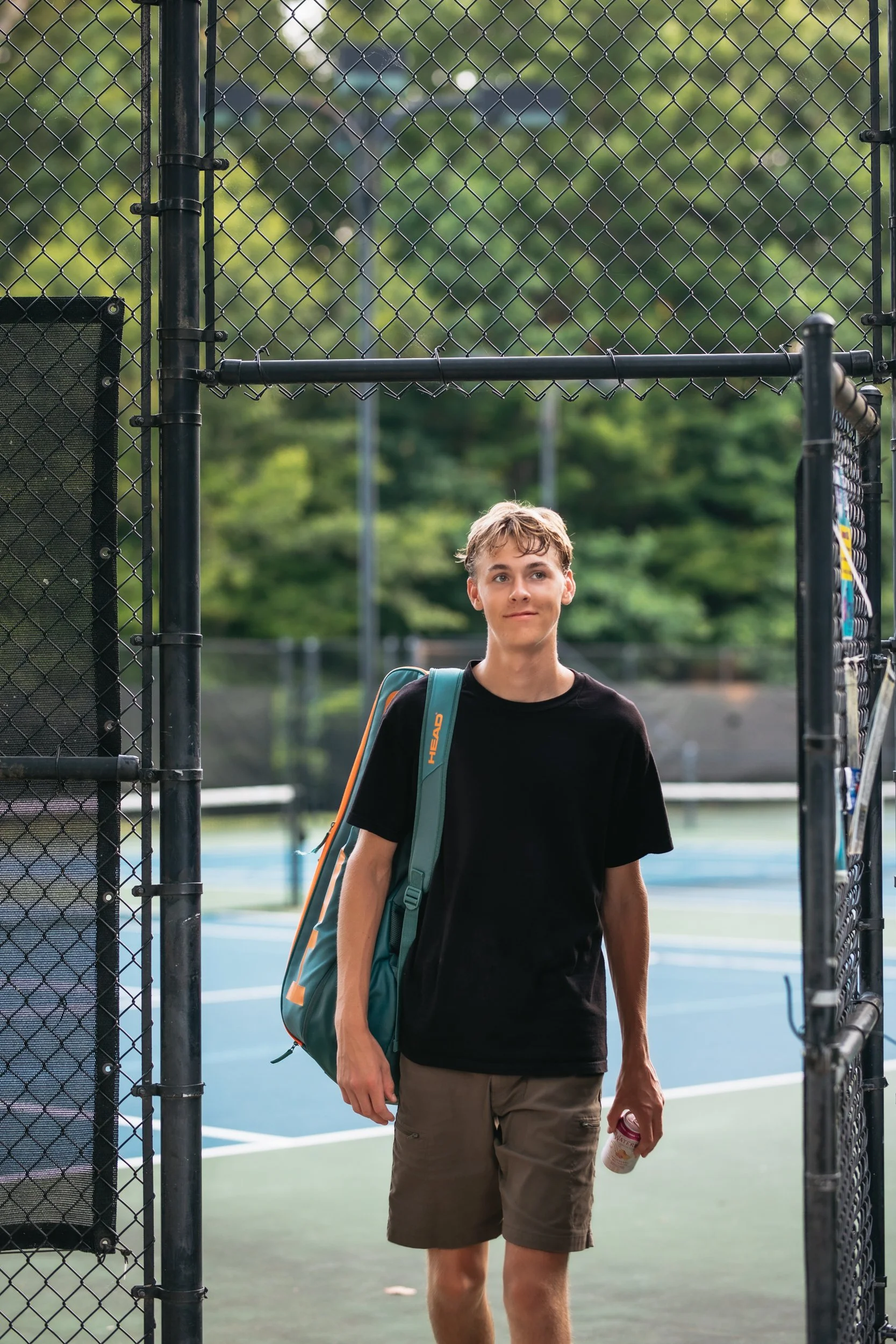A teenage boy with a backpack and a water bottle stands on a tennis court, framed by a chain-link fence, with trees in the background.