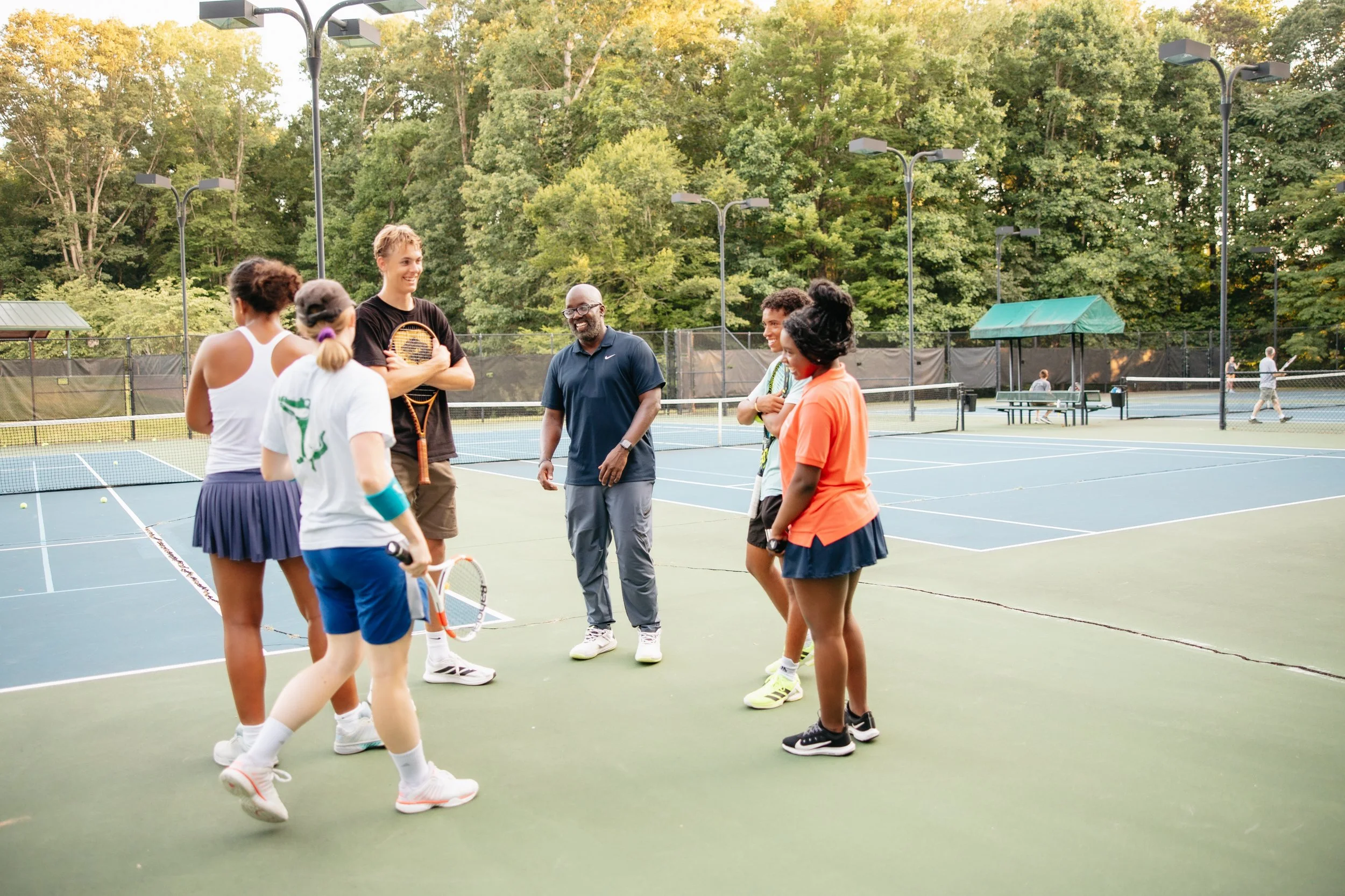 A group of six people, including men and women, standing on a tennis court, engaged in conversation, with tennis balls and rackets visible. The background shows trees and other tennis courts.