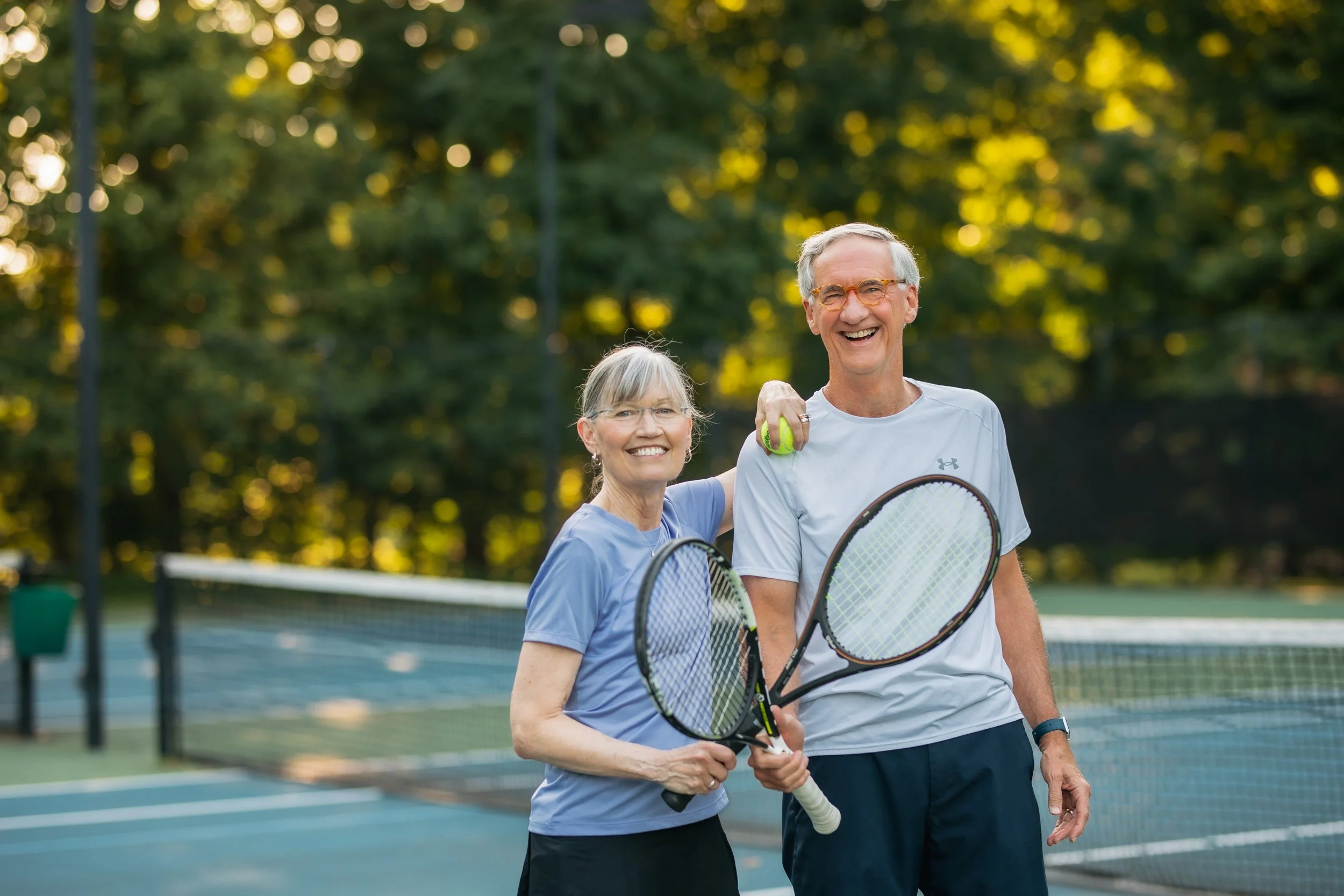 A smiling elderly couple stands side by side on a tennis court holding racquets and tennis balls in a sunny outdoor setting, surrounded by green.
