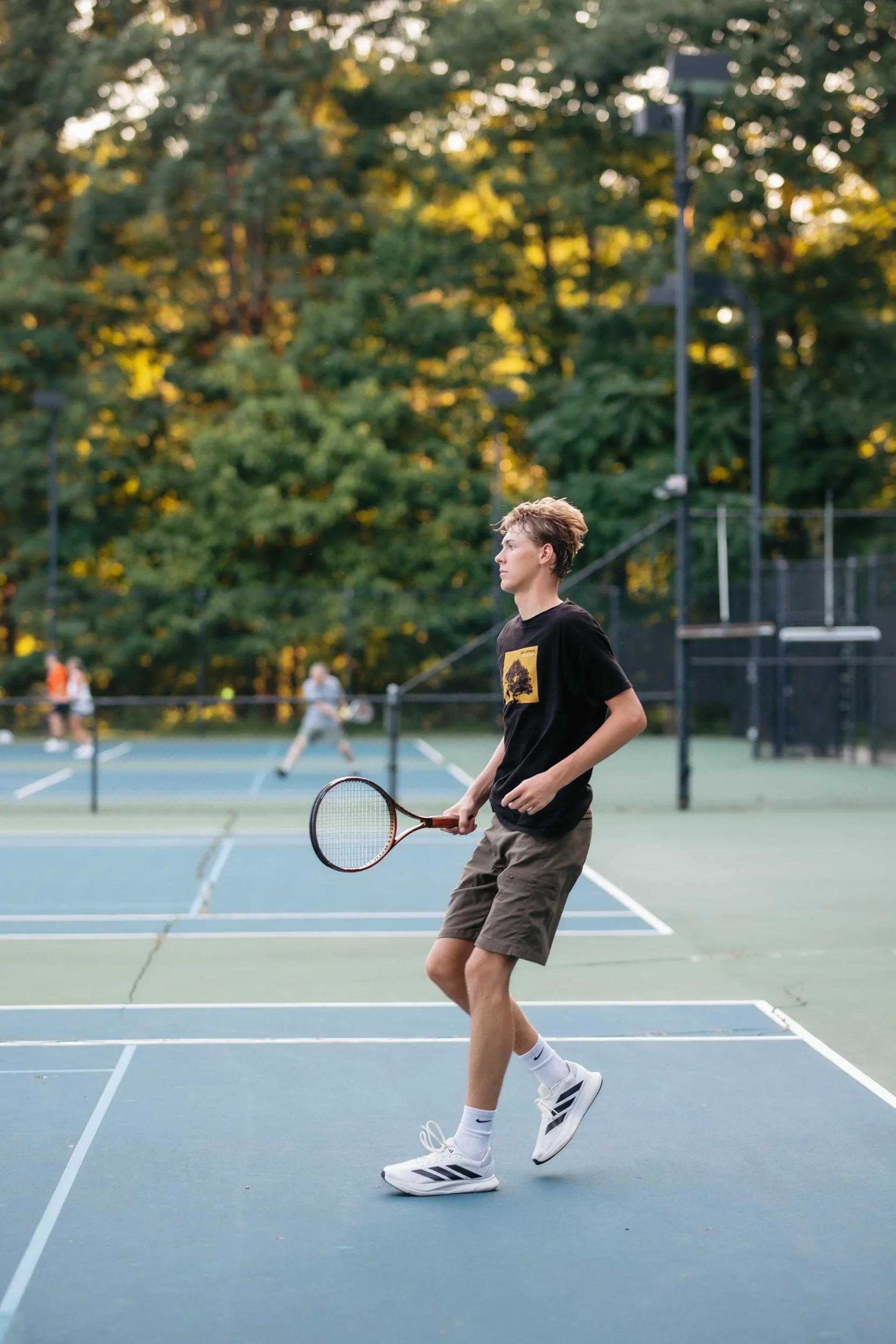 A young man playing tennis on an outdoor court, holding a racket, with a net and other players in the background, surrounded by trees.