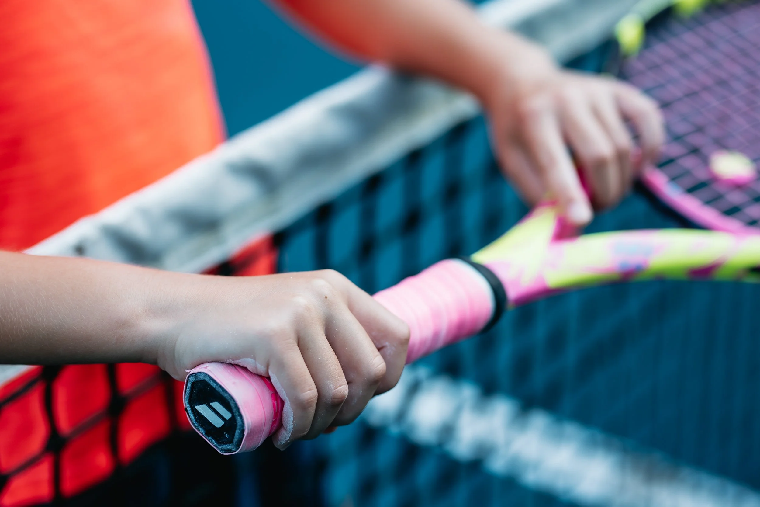 Close-up of a child's hand gripping a pink tennis racquet on a tennis court with a person in an orange shirt in the background.