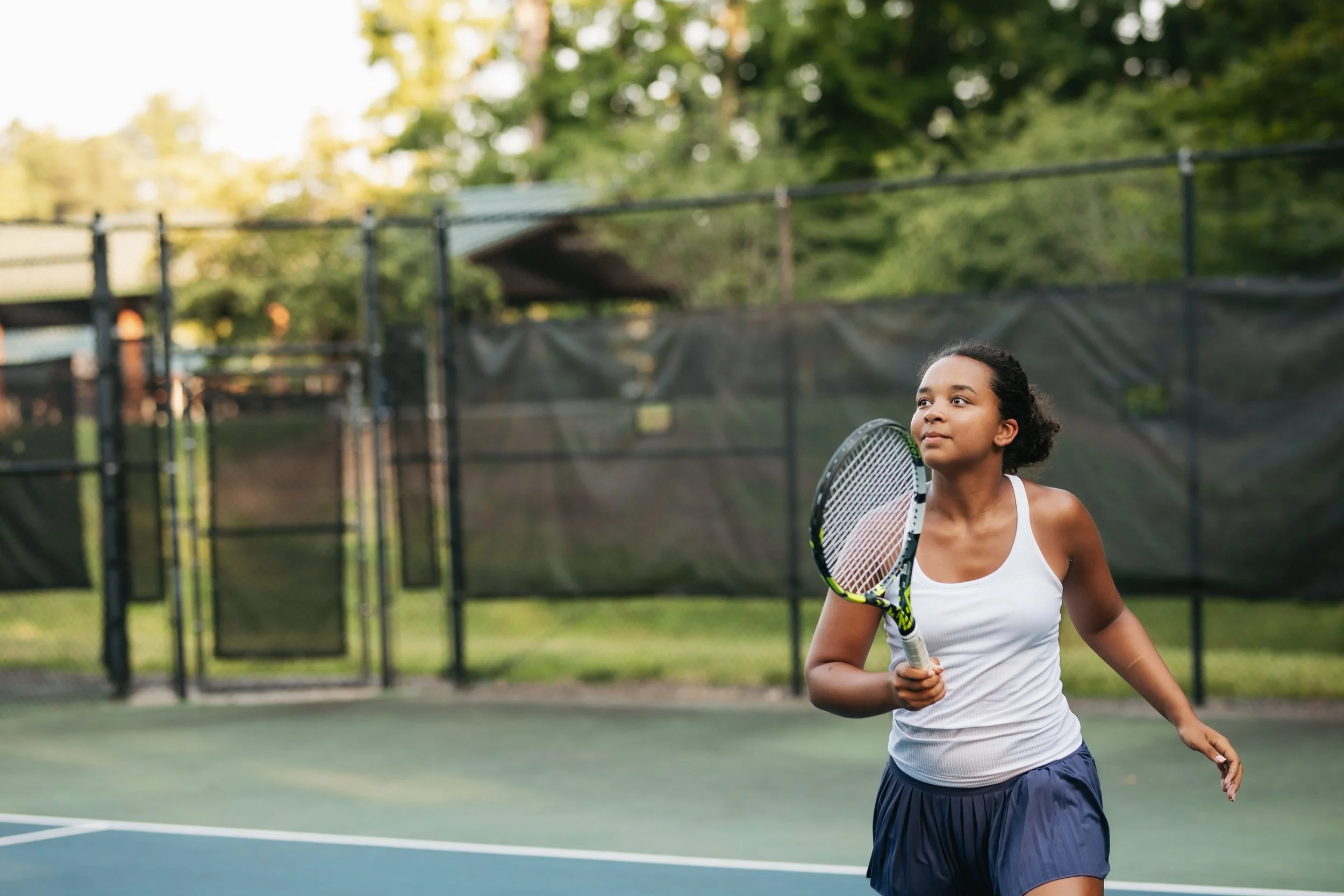 Young woman playing tennis on an outdoor court, holding a tennis racket, wearing a white tank top and navy skirt.