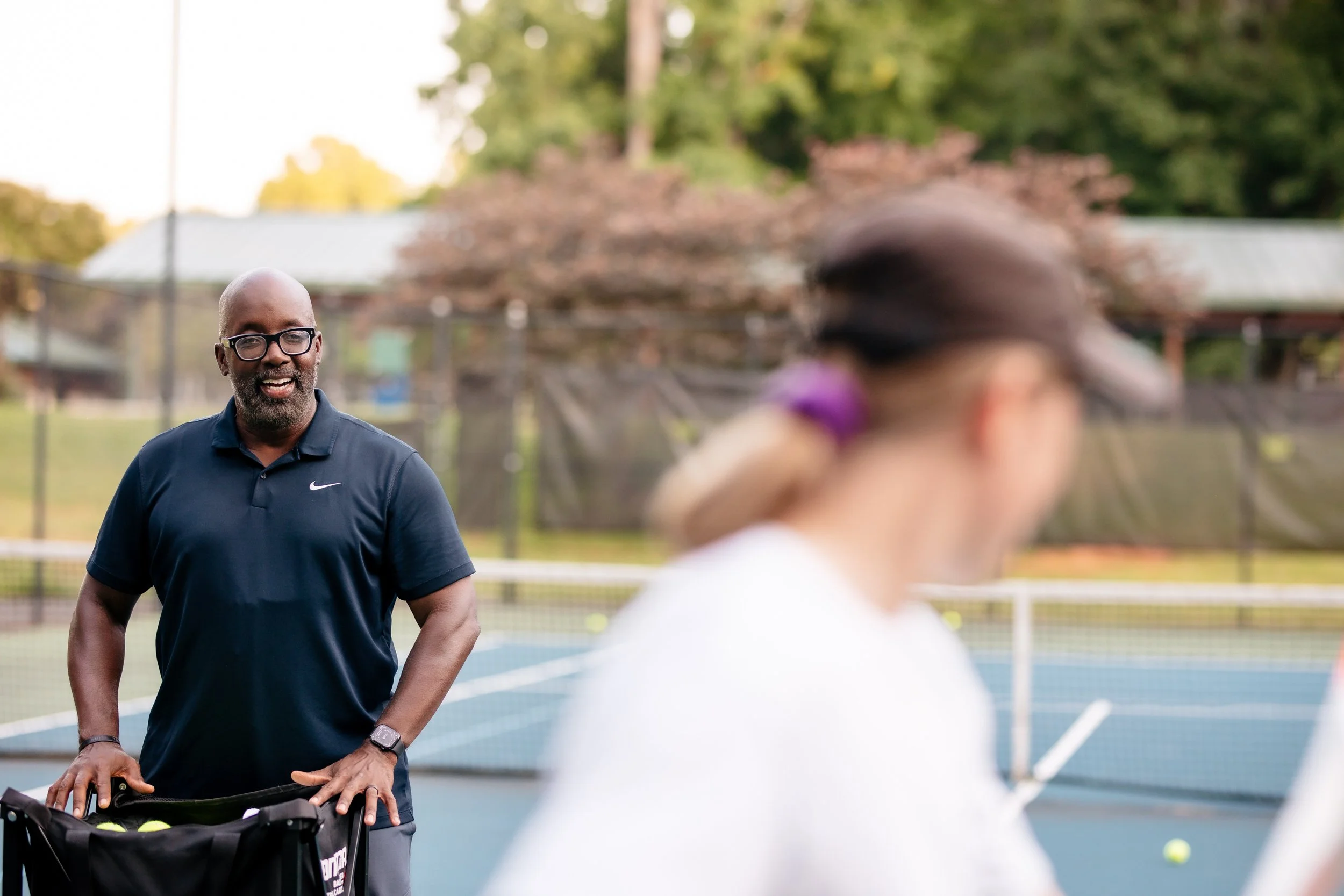 Owner of Triad Tennis, Denard McLendon sports glasses and a beard and smiles on a tennis court, the blurred form of a woman in a baseball cap standing in the foreground holding a tennis racquet.