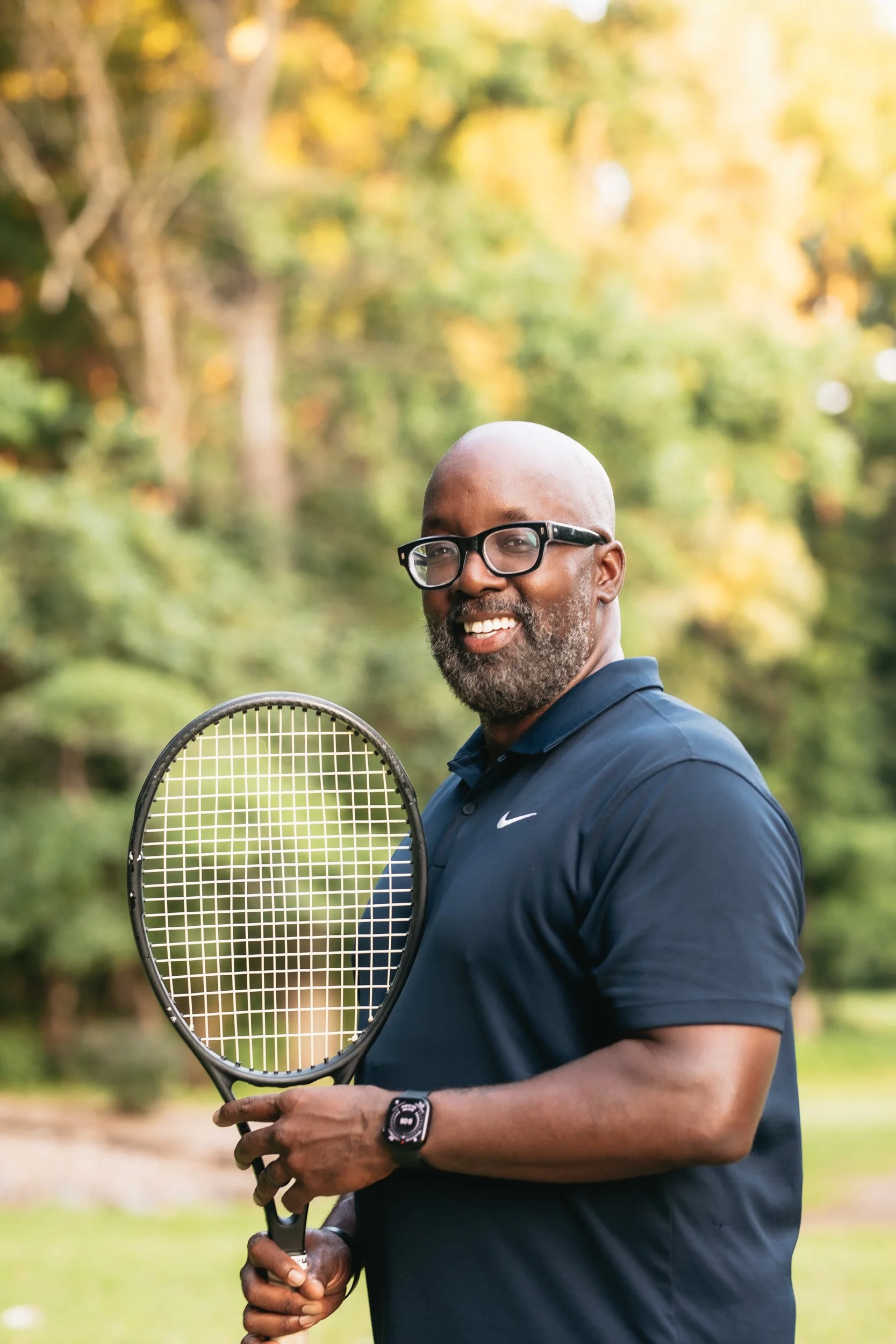 A smiling man with glasses and a beard wearing a navy Nike polo shirt, holding a tennis racket, standing outdoors in a park with trees and autumn foliage in the background.
