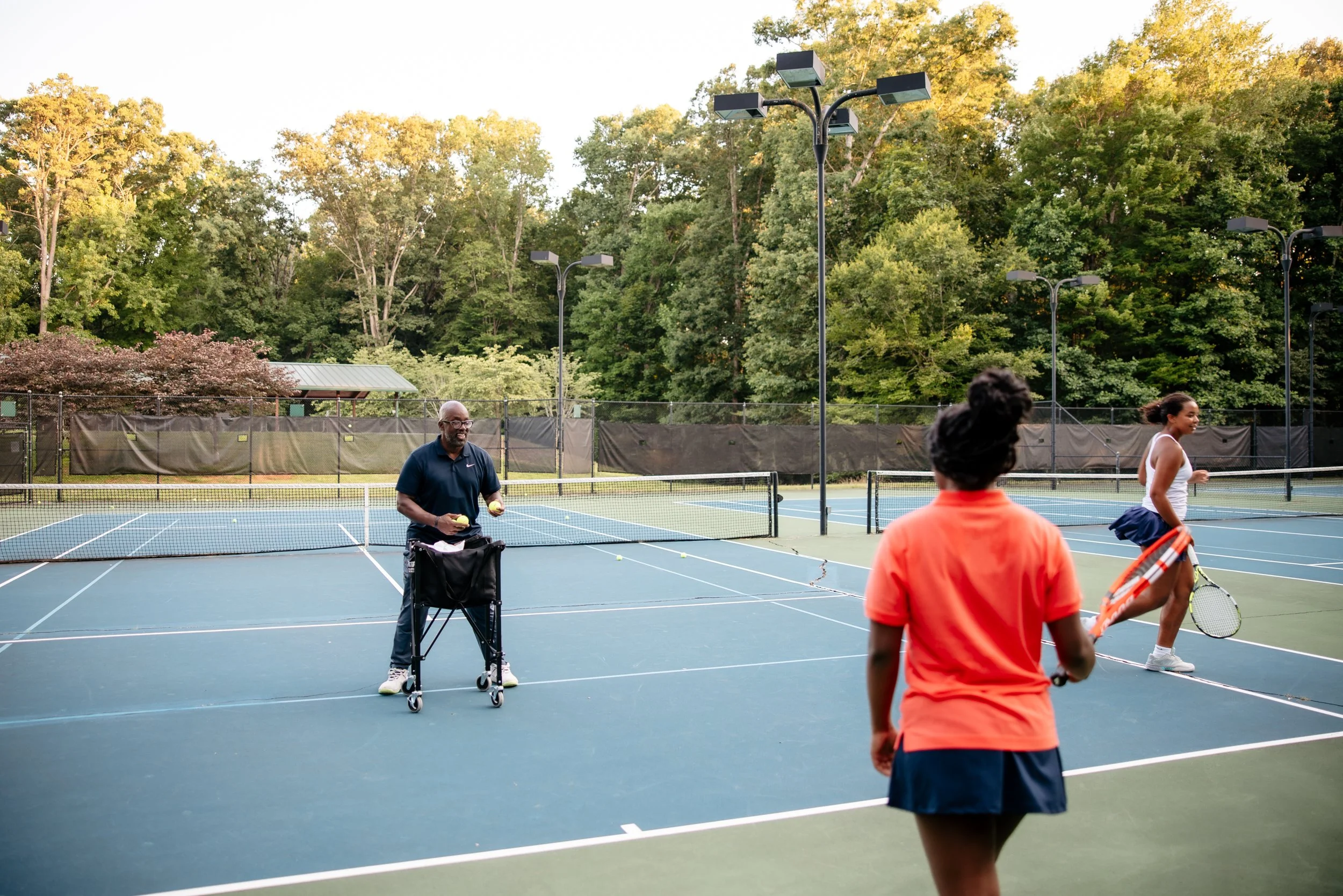 Three people playing tennis on an outdoor court surrounded by trees, with one person tossing tennis balls and two women holding rackets.