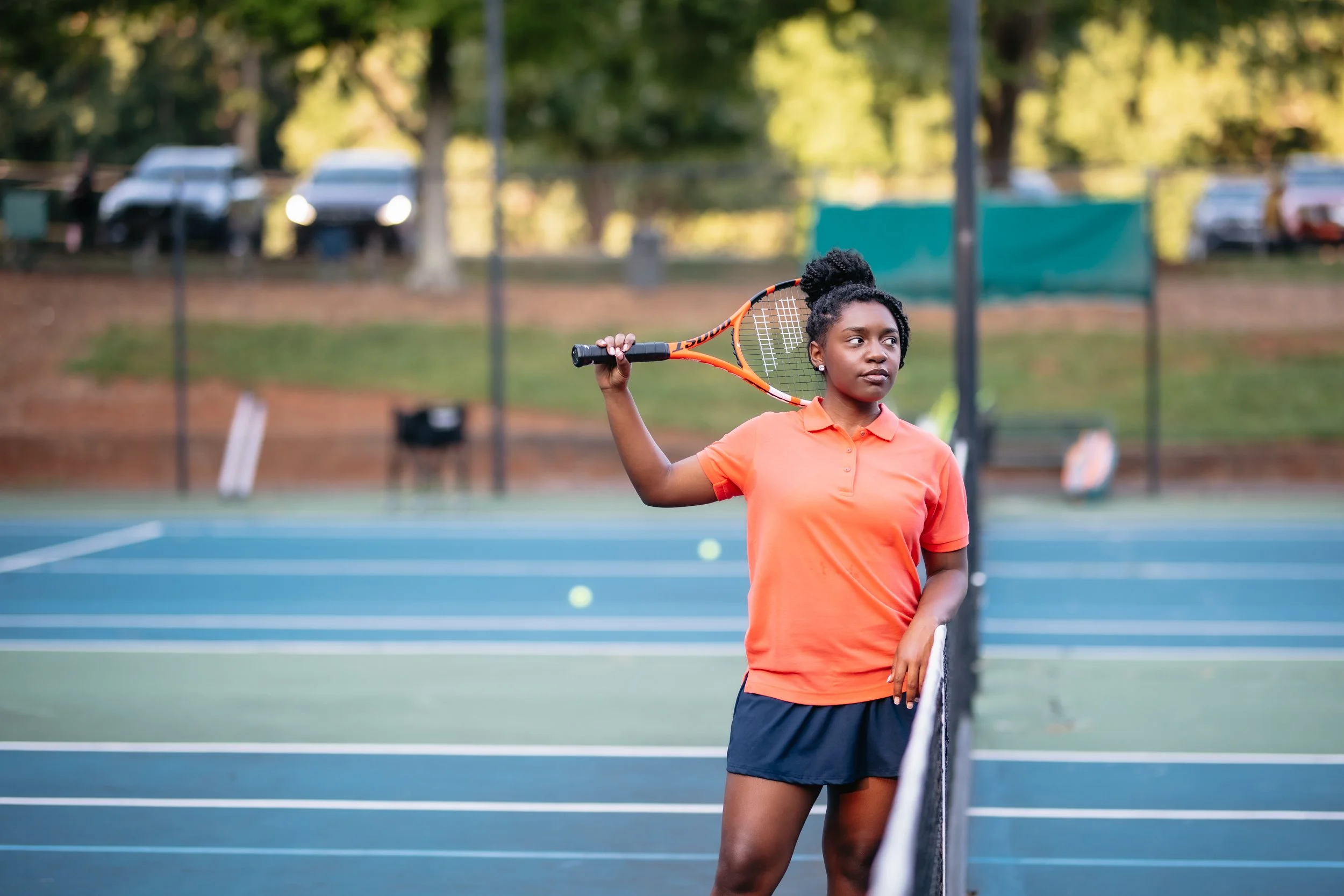 A young woman wearing an orange polo shirt and black shorts, standing on a tennis court holding a tennis racket over her shoulder, looking to the side.