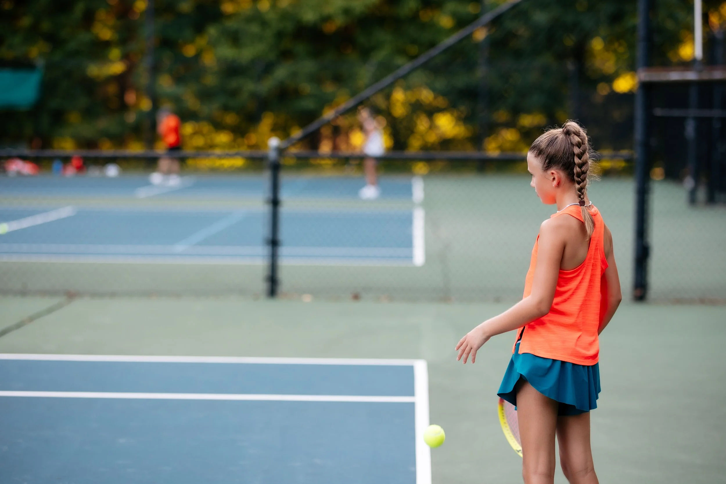 Young girl standing on a tennis court, holding a tennis racket, with a tennis ball nearby, and other players in the background.