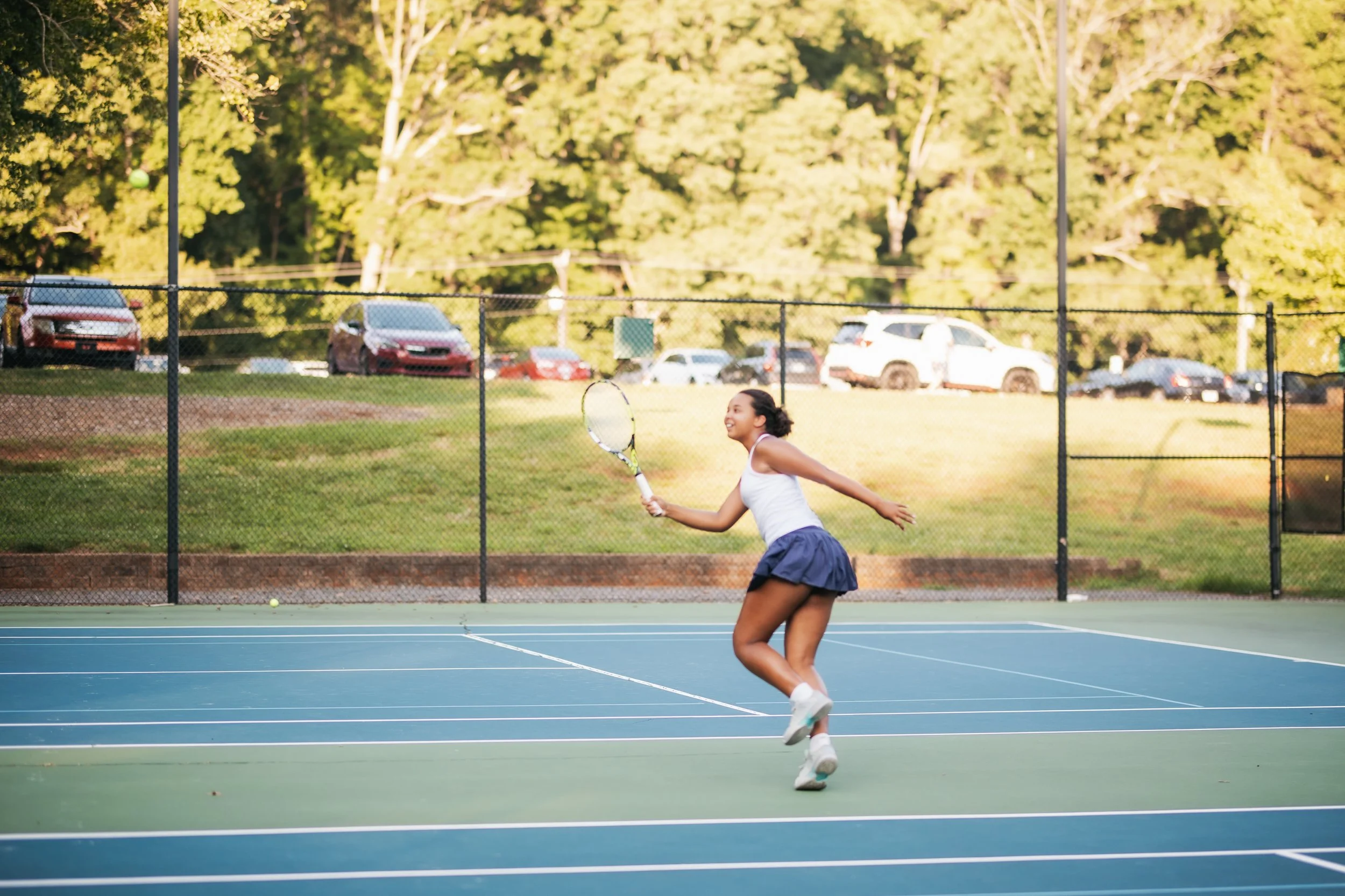 A young woman playing tennis on an outdoor court, smiling, with a tennis racket in her hand and a tennis ball nearby, surrounded by trees and parked cars.