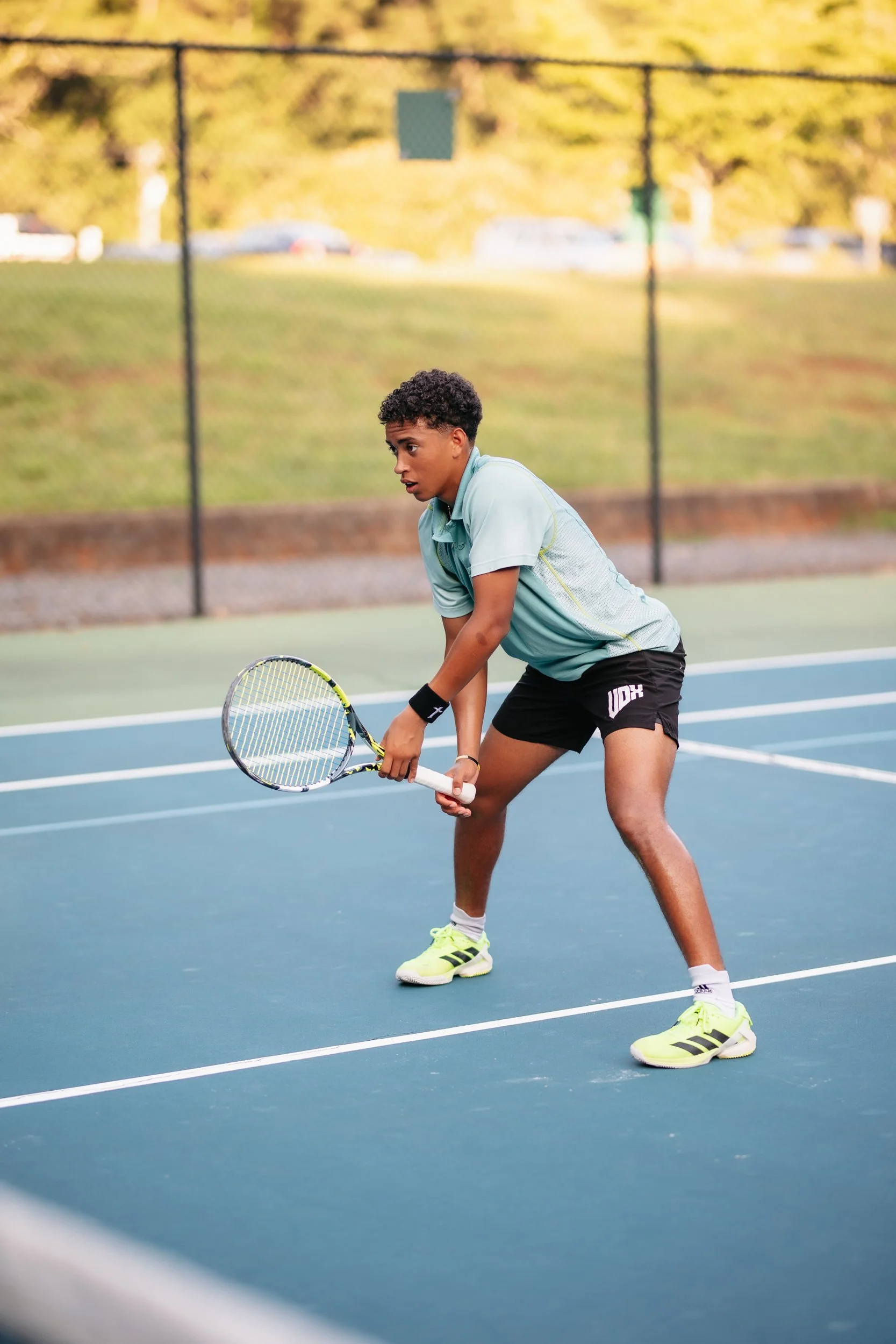 A young male tennis player dressed in a light blue shirt and black shorts, holding a tennis racket, on an outdoor tennis court during daytime.