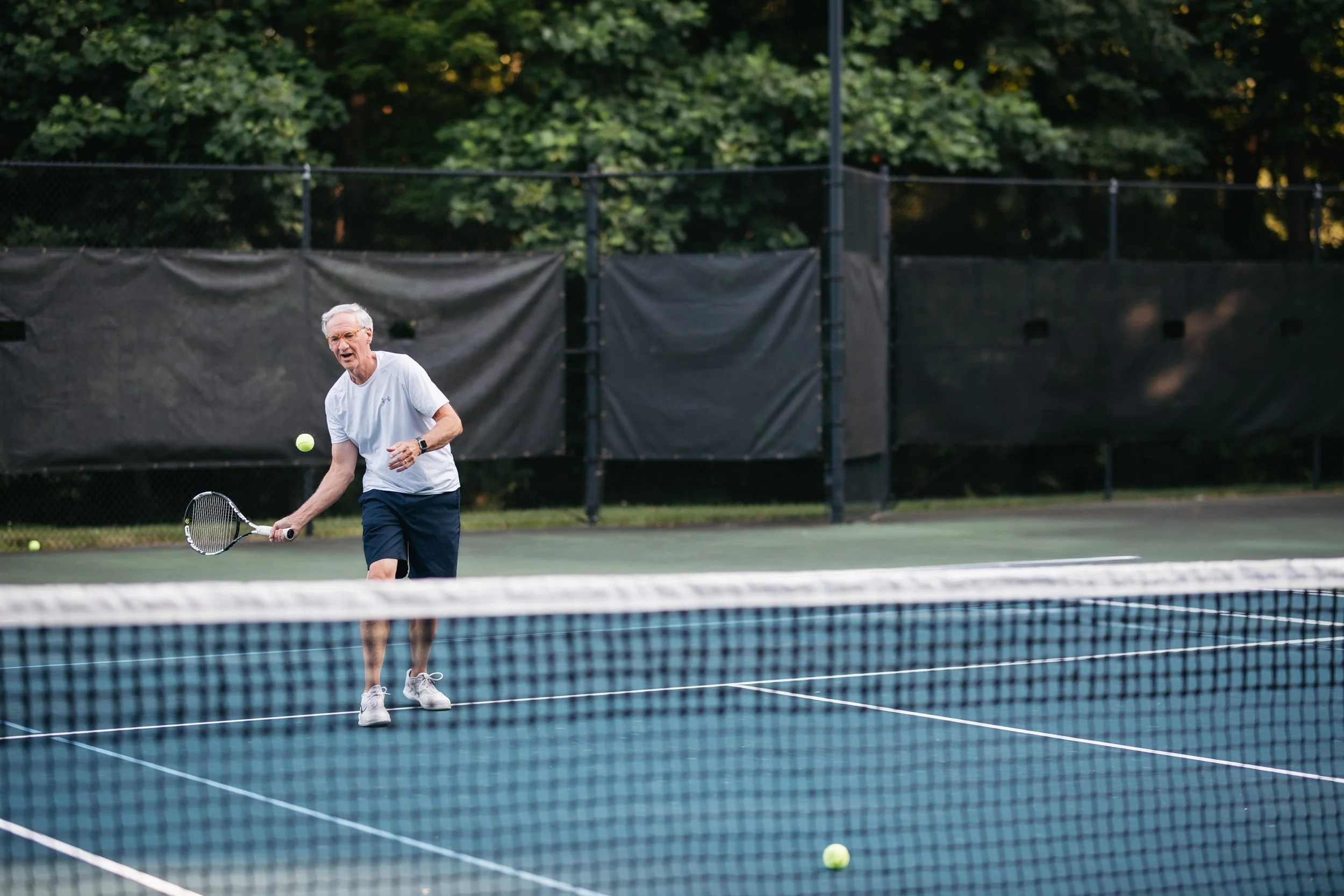 Older man playing tennis on an outdoor court, preparing to hit a tennis ball with a racket, with trees in the background.