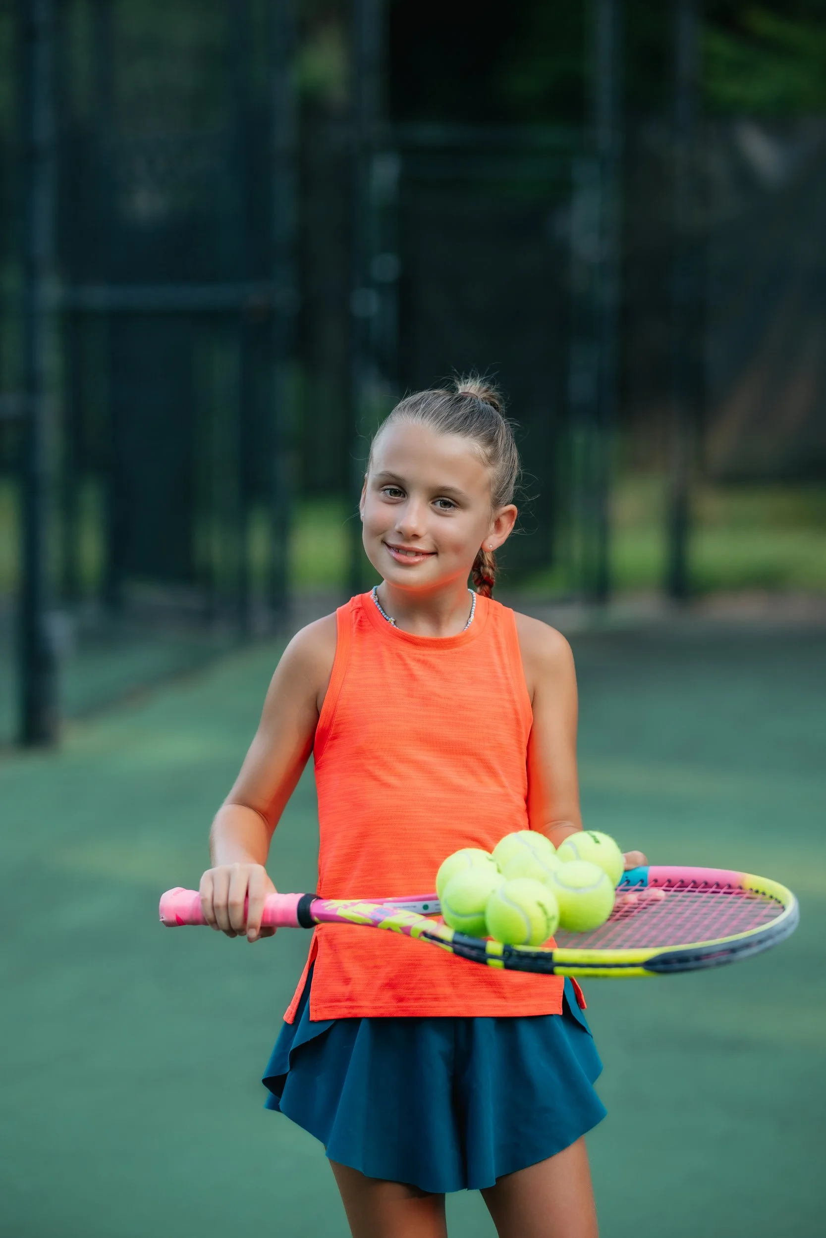 Young girl in orange tank top and navy shorts holding tennis racket with tennis balls on it, standing on a tennis court.