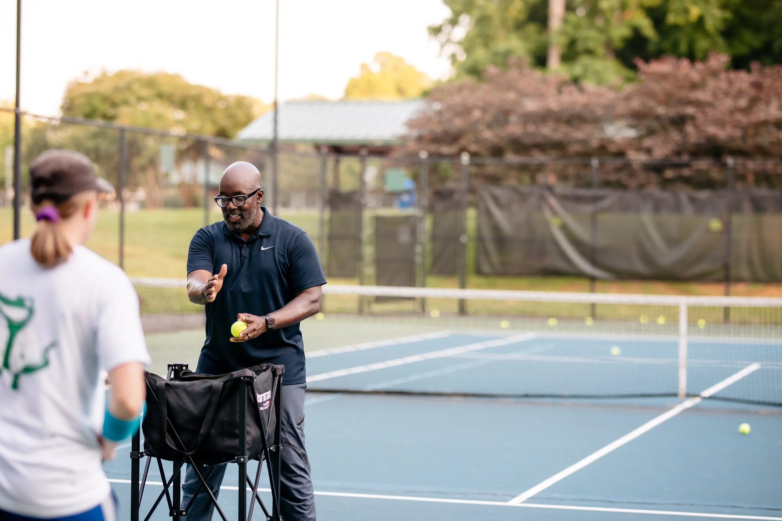 A tennis coach teaching a young girl on an outdoor tennis court with tennis balls scattered around.
