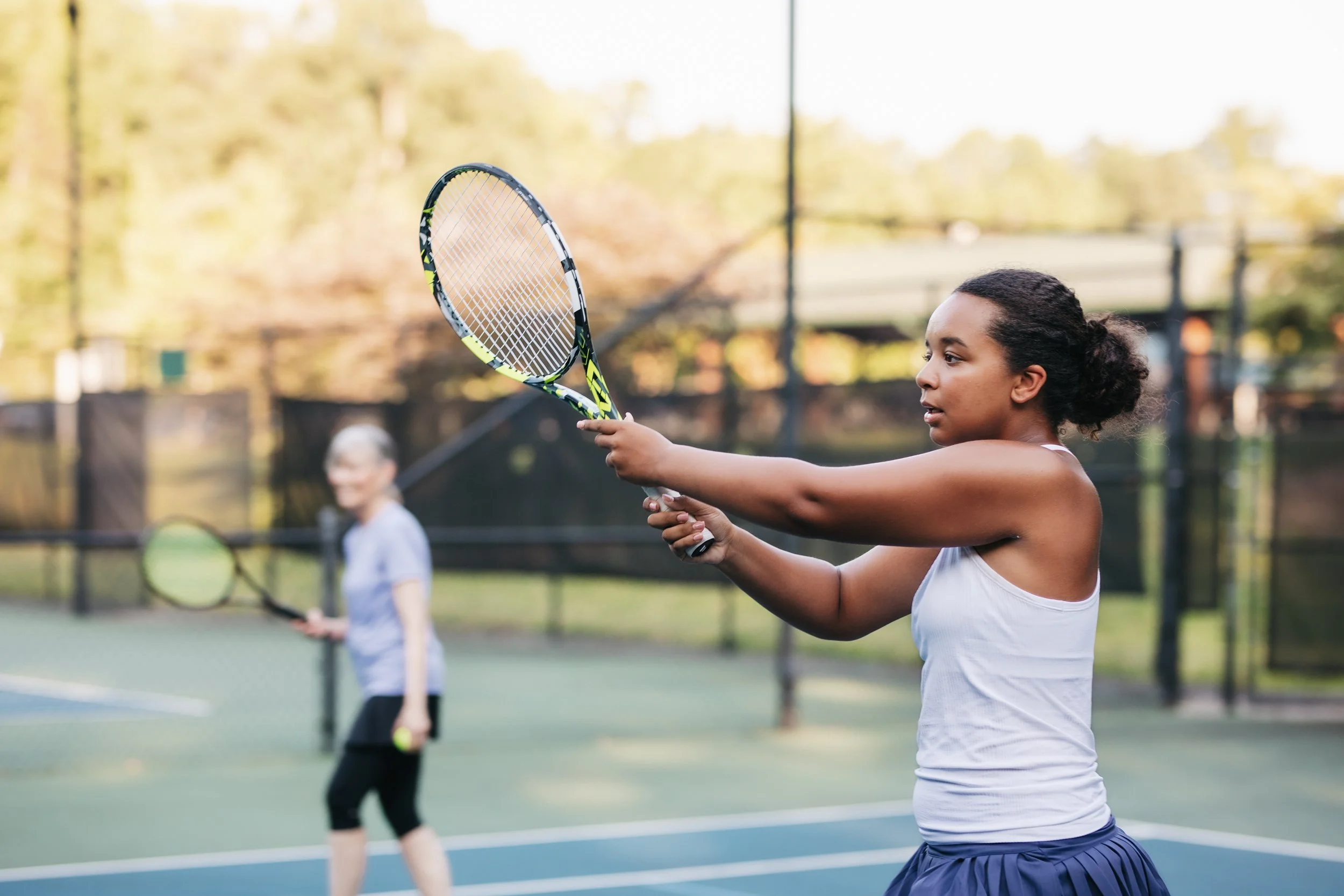 Two women are playing tennis on an outdoor court, one in the foreground preparing to hit the ball and one in the background holding a tennis racquet.
