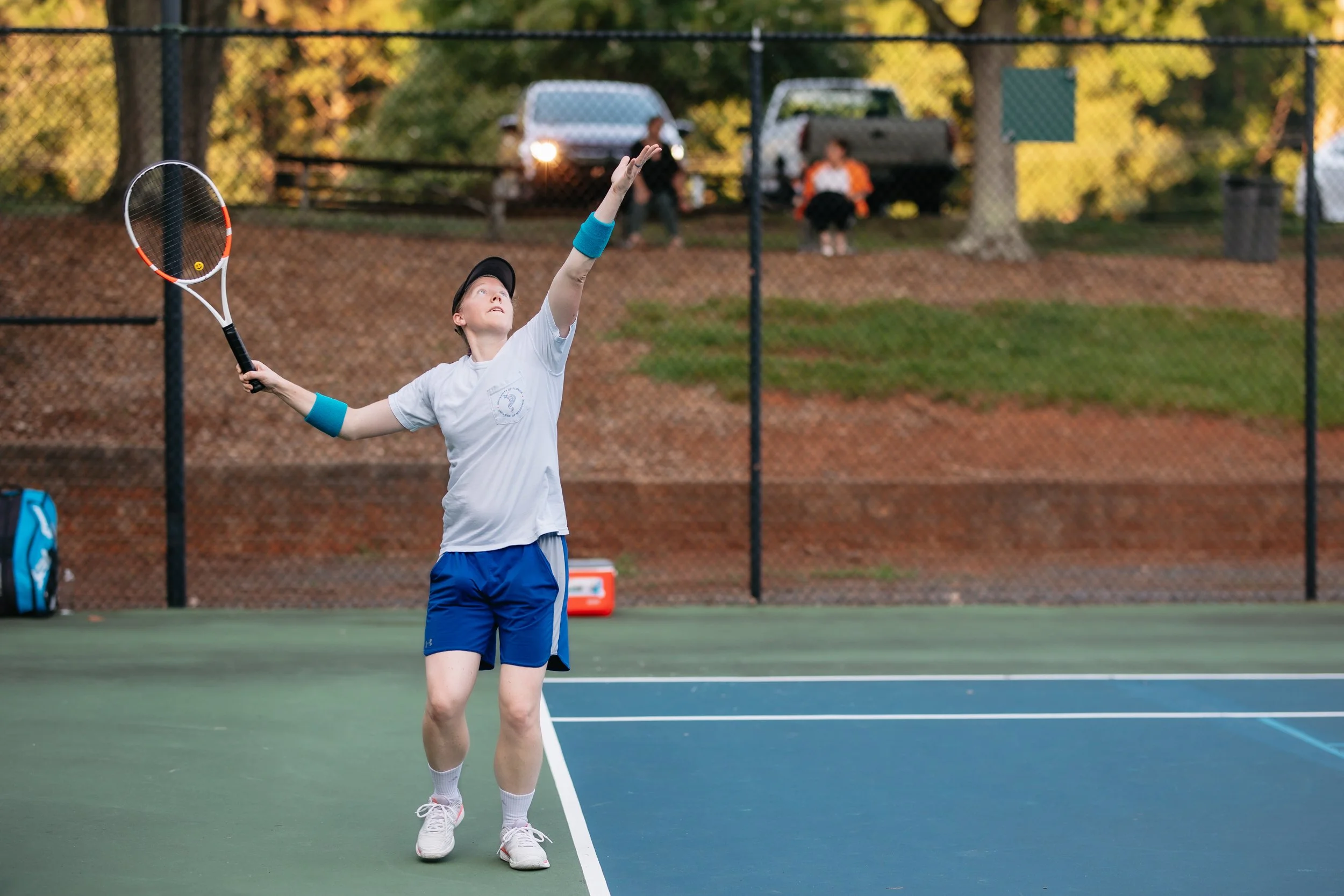 A young tennis player in a white shirt, blue shorts, and a black cap serving on an outdoor tennis court. The player has blue wristbands on both wrists and is holding a tennis racket, preparing to hit the ball. There are cars and spectators in the bac