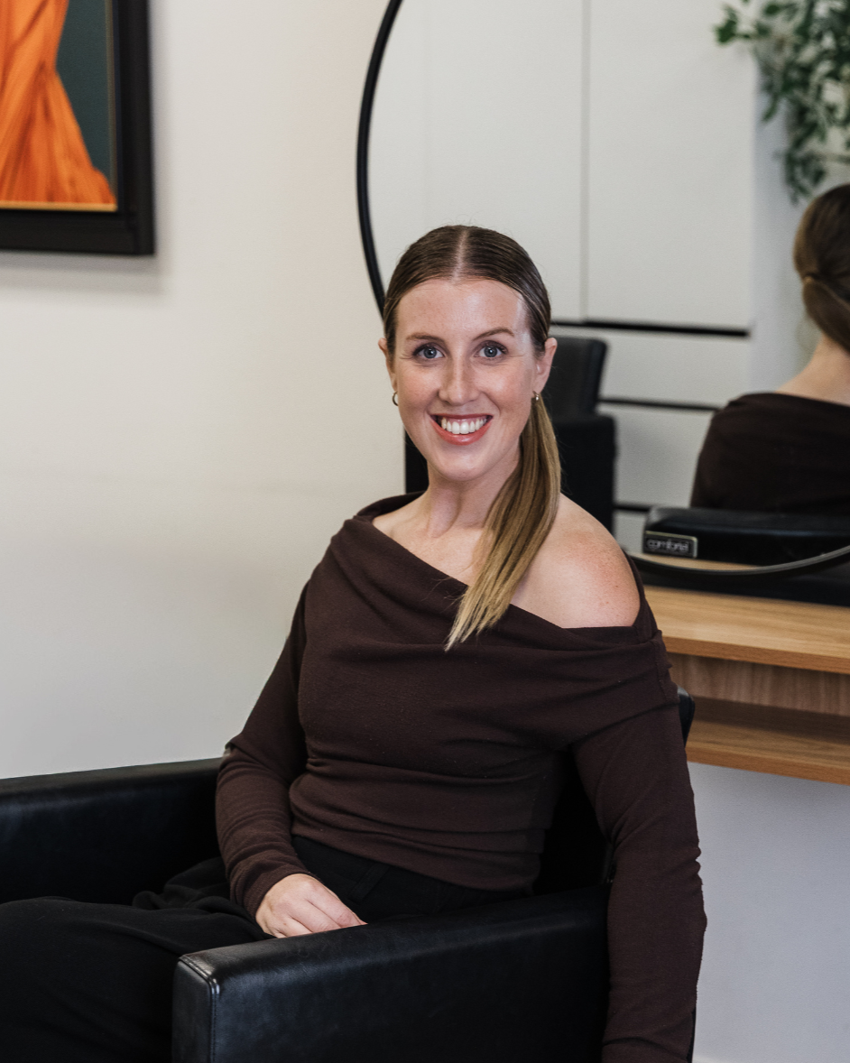 A woman with long blonde hair, blue eyes, and a nose piercing, sitting in a black chair, smiling at the camera in a modern salon or hair studio.