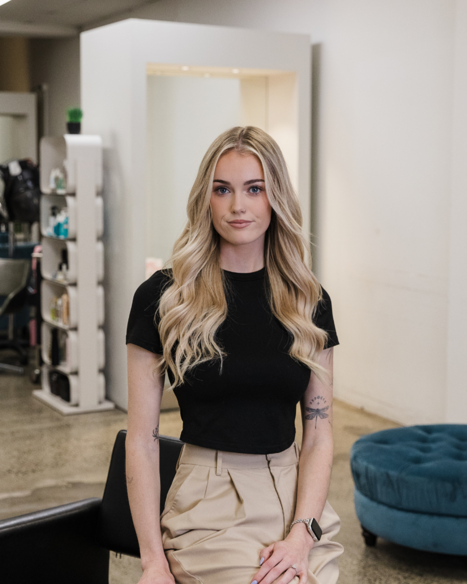 A young woman with long blonde wavy hair, blue eyes, and wearing a black and white checkered shirt sitting in a salon chair.