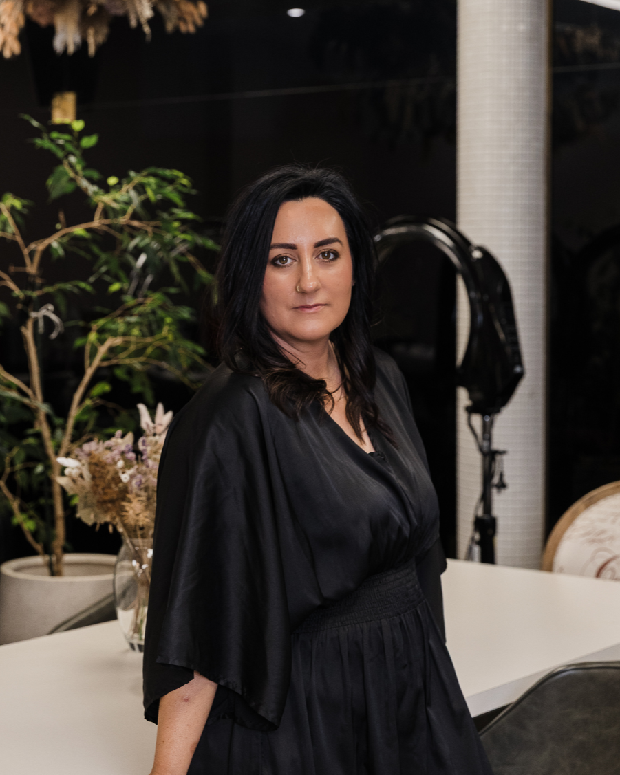 A woman with long black and brown hair, wearing a black dress, sitting at a white table in a modern, well-lit office or salon environment with chairs and plants in the background.
