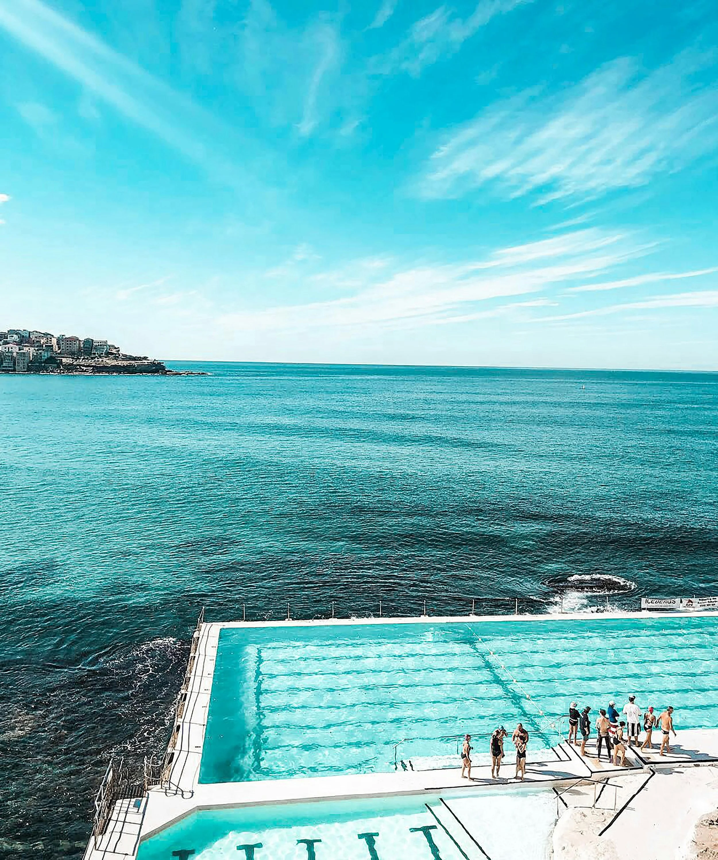 People gather around an outdoor swimming pool overlooking the ocean with a coastal city in the distance under a partly cloudy sky.