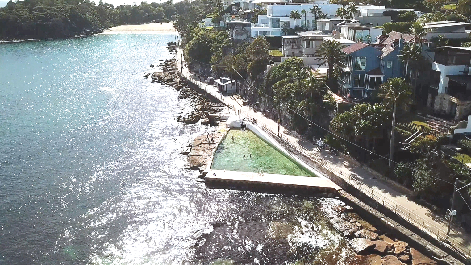 Coastal scene with a swimming pool on the rocky shoreline, adjacent to a walkway and residential houses on a hillside near the ocean.