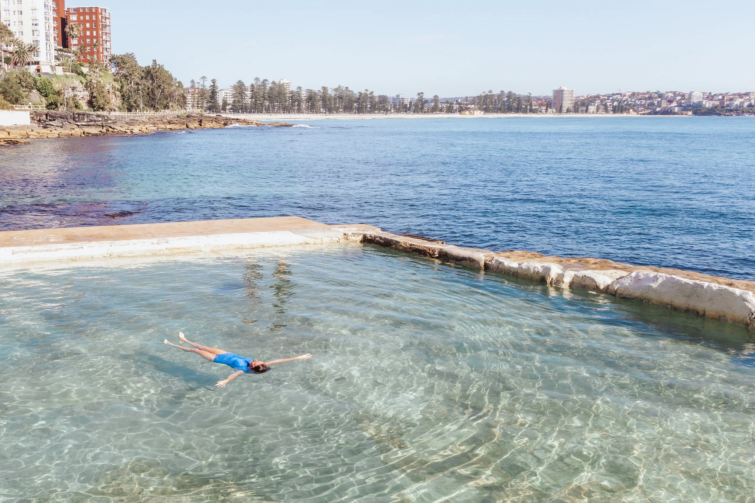 A person floating on their back in a clear, shallow outdoor pool near the shoreline with city buildings in the background.