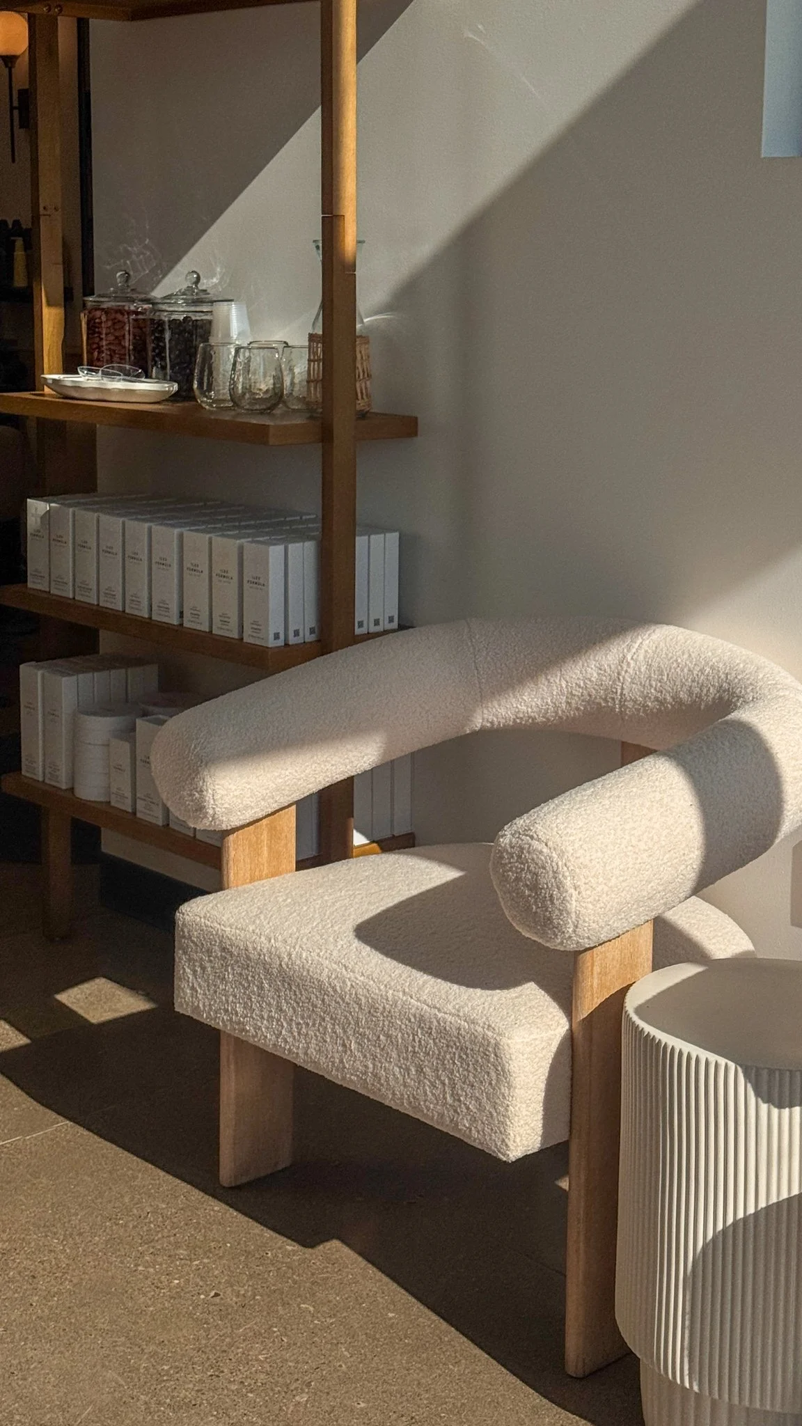 A modern cream-colored armchair with rounded armrests placed next to a white textured side table, with a wooden shelf behind displaying glass jars filled with beans, cups, and boxes.