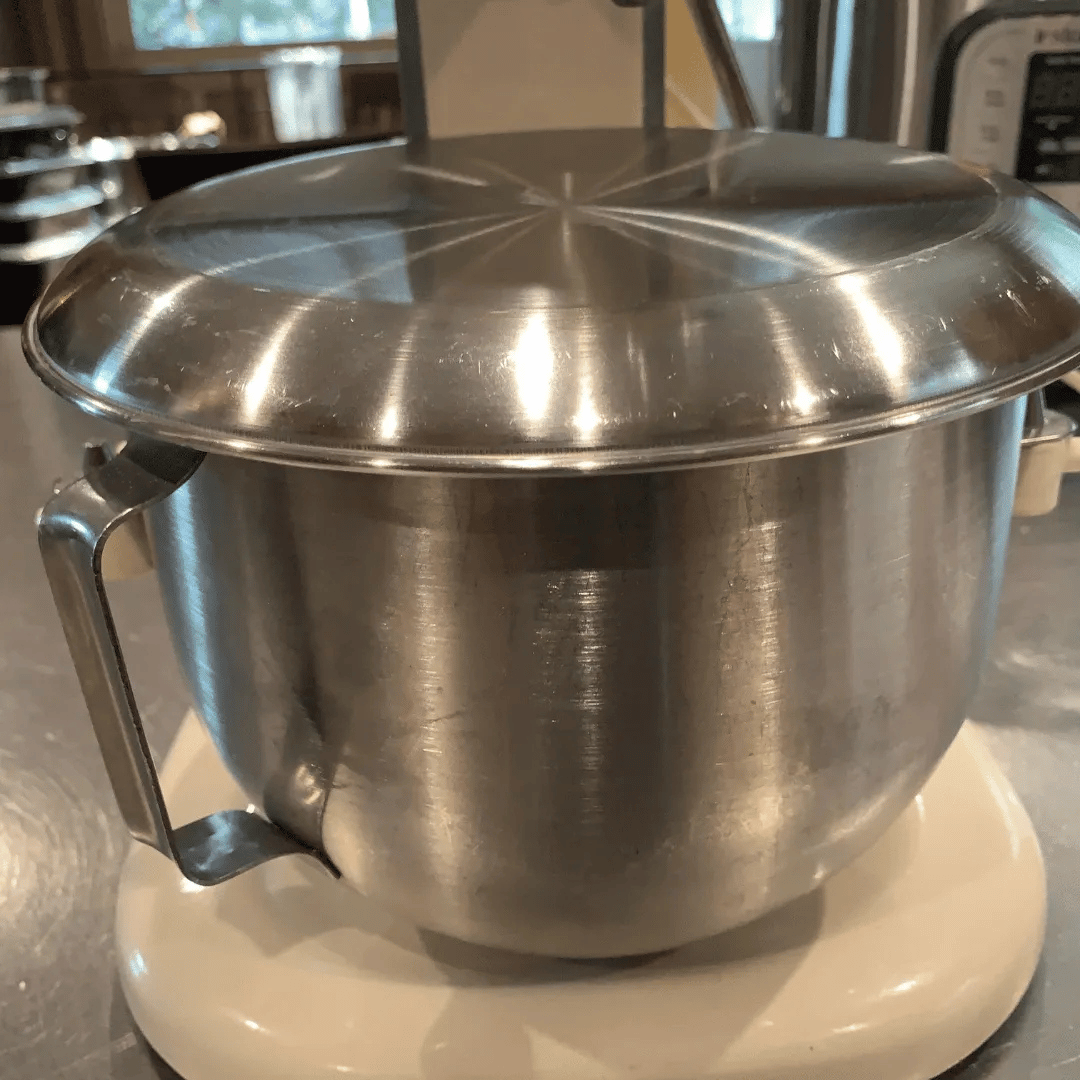 Stainless steel mixing bowl with a lid, sitting on a white scale in a kitchen setting.