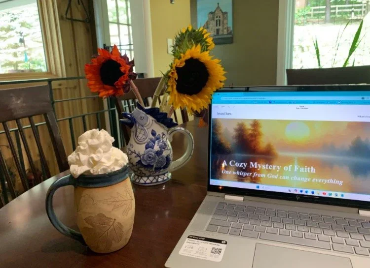A laptop on a wooden table displaying a website with the title "A Cozy Mystery of Faith." Next to the laptop is a mug filled with whipped cream, a decorative blue and white pitcher holding sunflowers, and a vase with red and yellow sunflowers. The background shows a bright dining room with windows and a door, with some outdoor scenery visible.