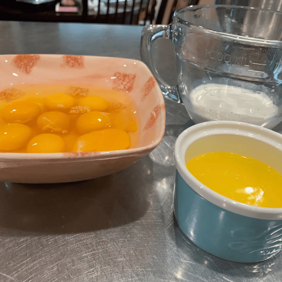 A bowl of raw eggs, a measuring cup with flour, a small container with more eggs, and a glass measuring cup with water on a stainless steel countertop.