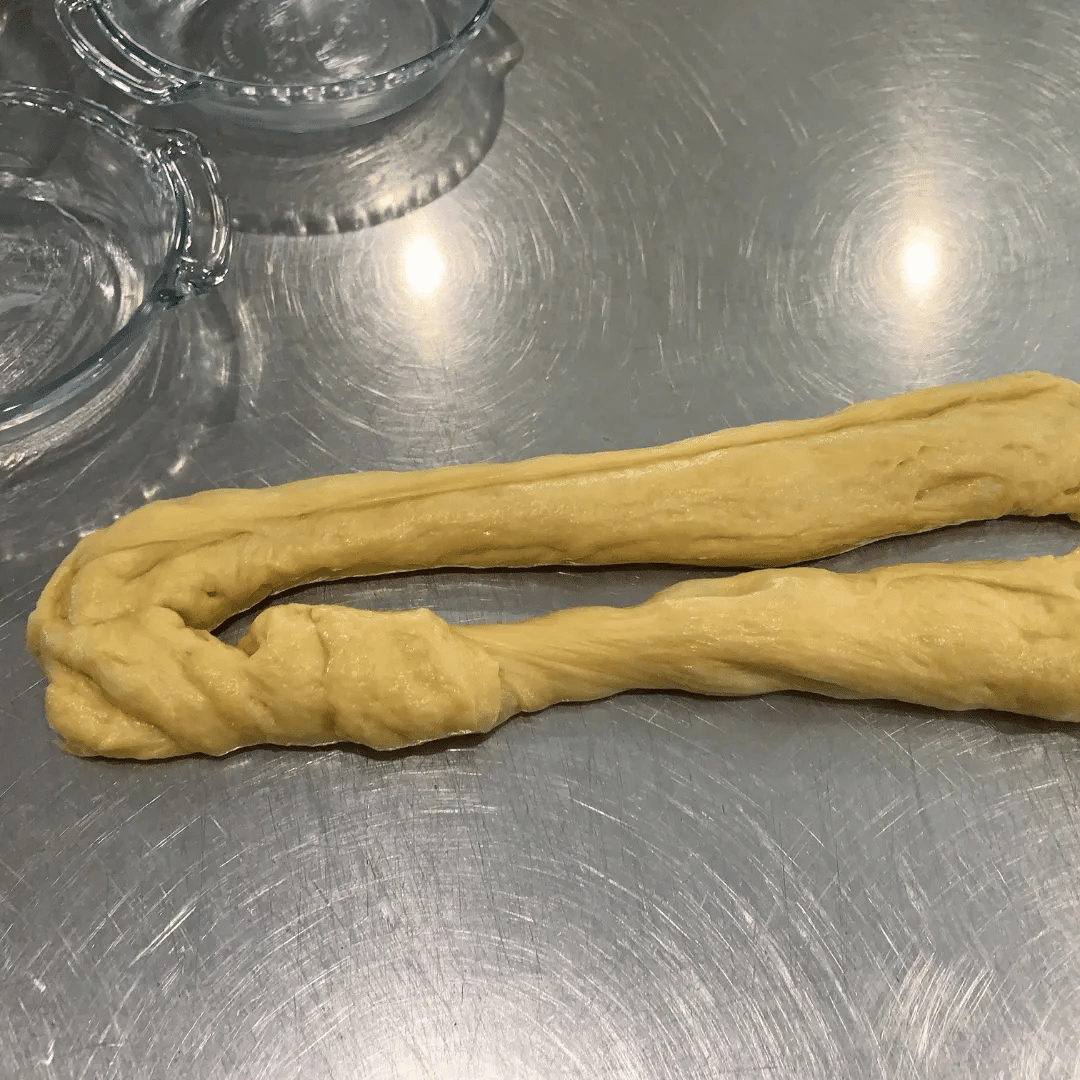 Two pieces of twisted dough on a metal surface with two glass bowls nearby.