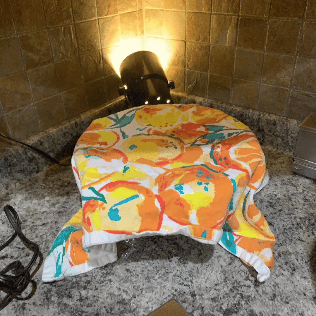 A cloth with a colorful orange, yellow, and teal fruit pattern draped over a plate on a granite countertop in a kitchen.
