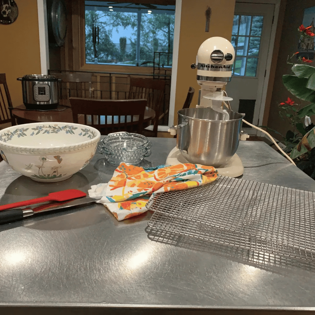 Kitchen countertop with mixing bowl, glass bowl, kitchen aid stand mixer, cooling rack, and dish towel, with a window and dining area in the background.