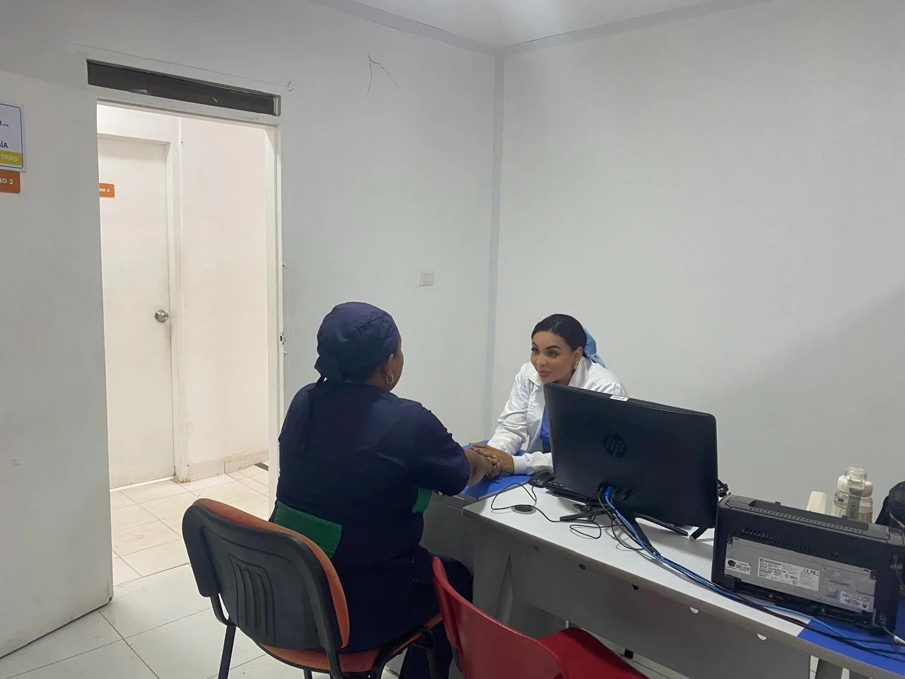 Una mujer en uniforme médico atendiendo a una paciente en consulta en un consultorio médico con escritorio, computadora y medicamentos en el escritorio.